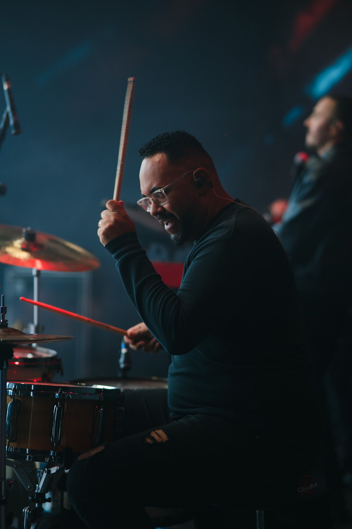 Baterista tocando bateria no palco, iluminado com luzes azuis e vermelhas.