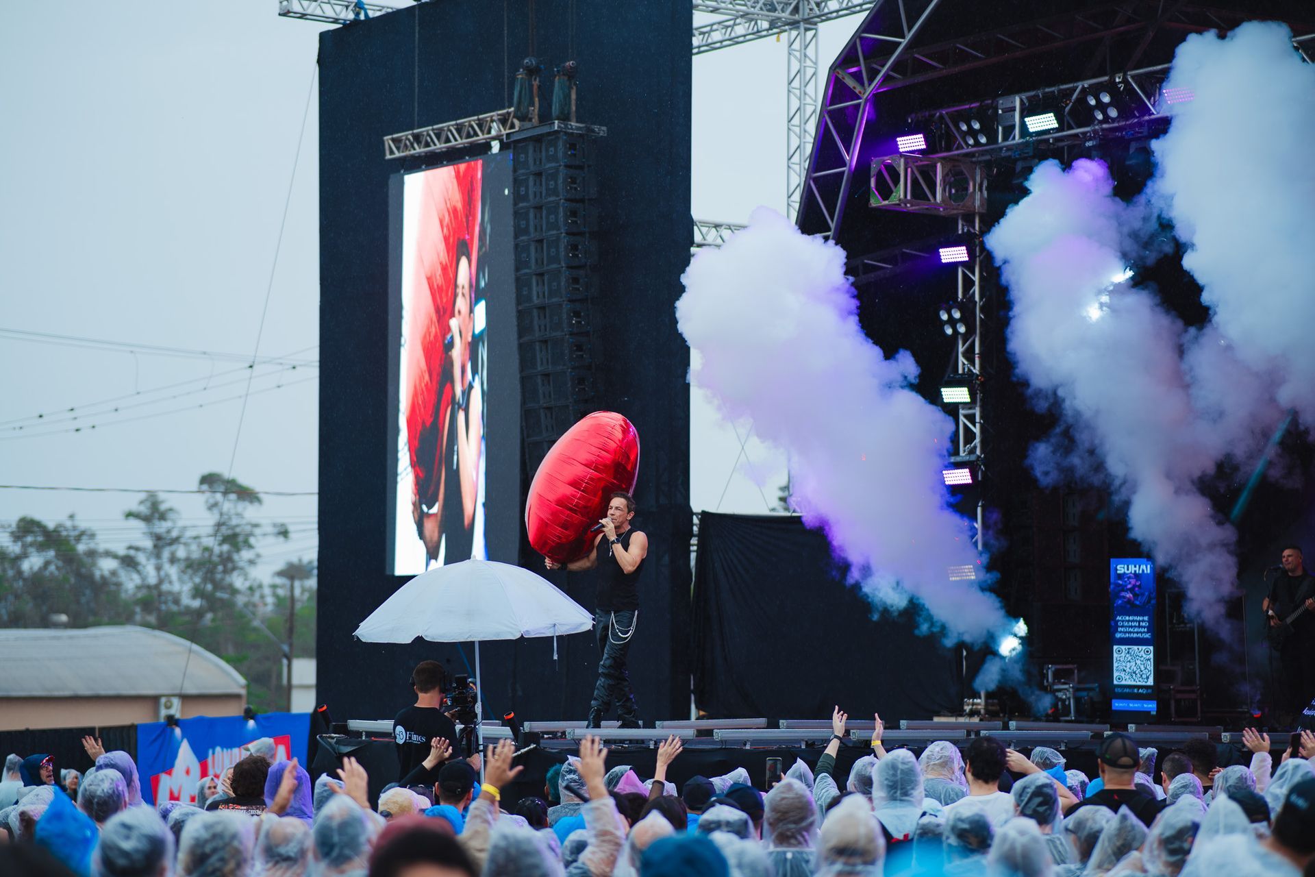 Um artista canta no palco; fumaça e uma multidão com capas de chuva são visíveis.