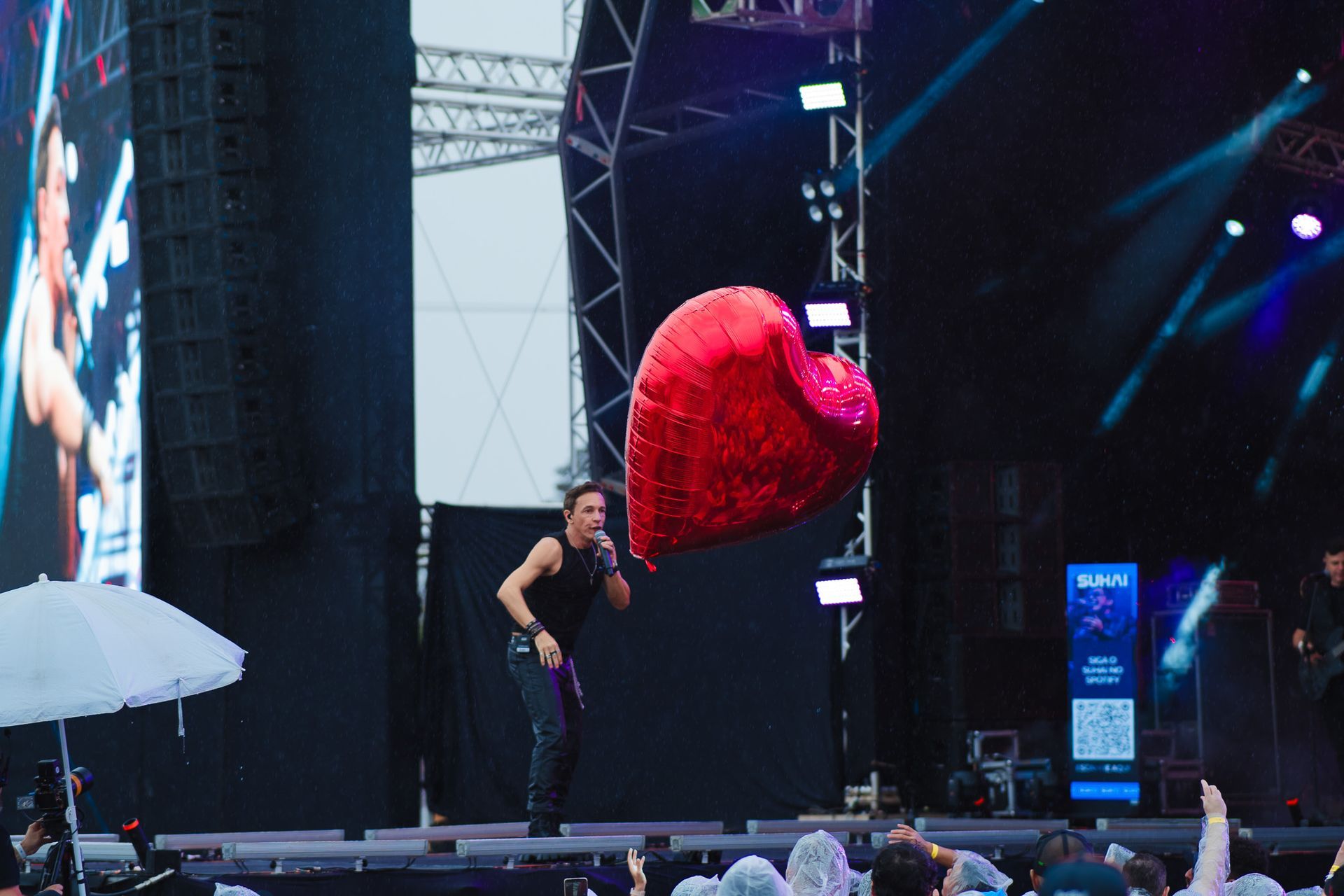 Cantor no palco com um balão vermelho em forma de coração, sob luzes de palco.