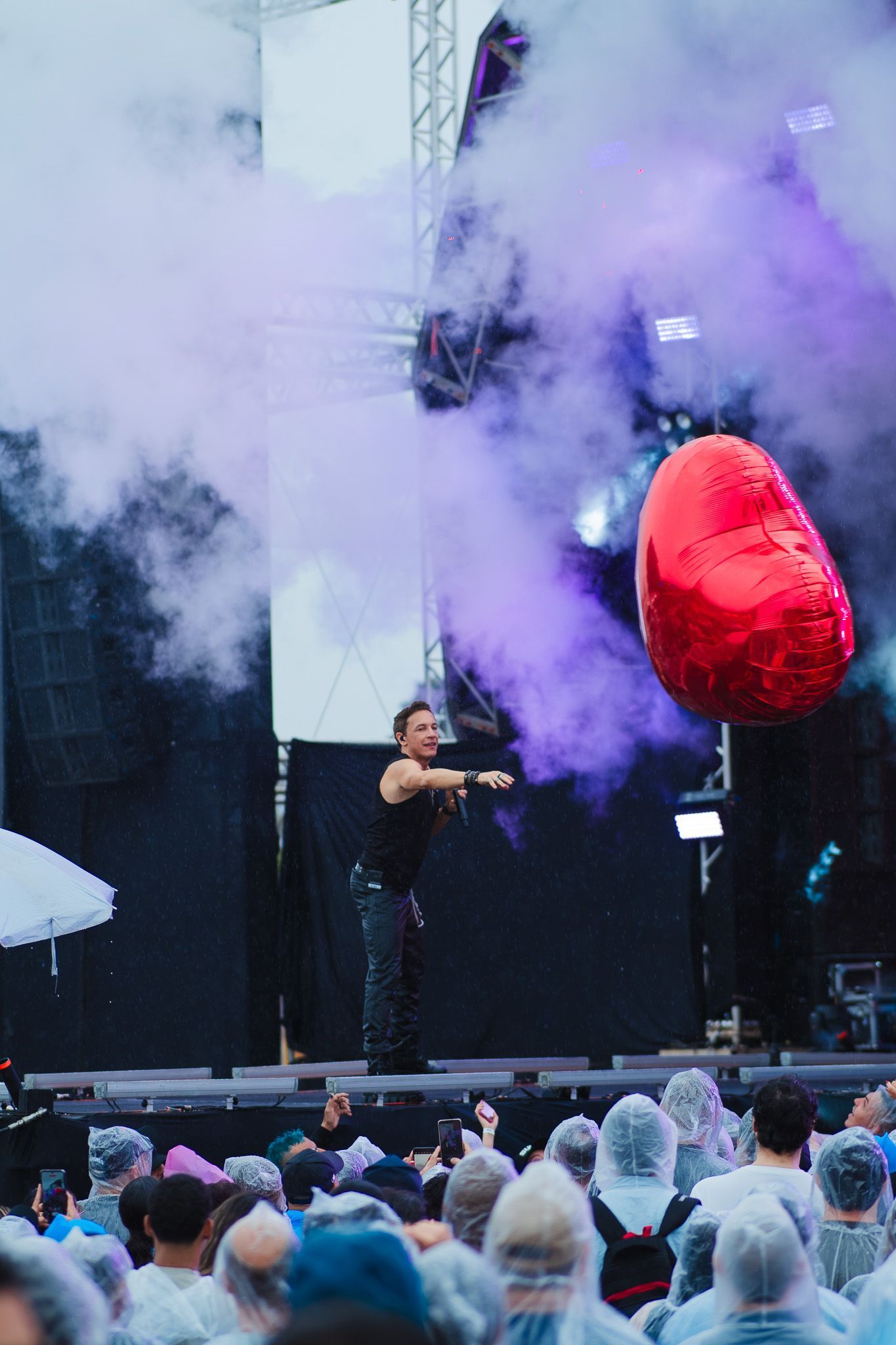 Um artista no palco, braços estendidos, perto de um balão vermelho em forma de coração, público com capas de chuva.