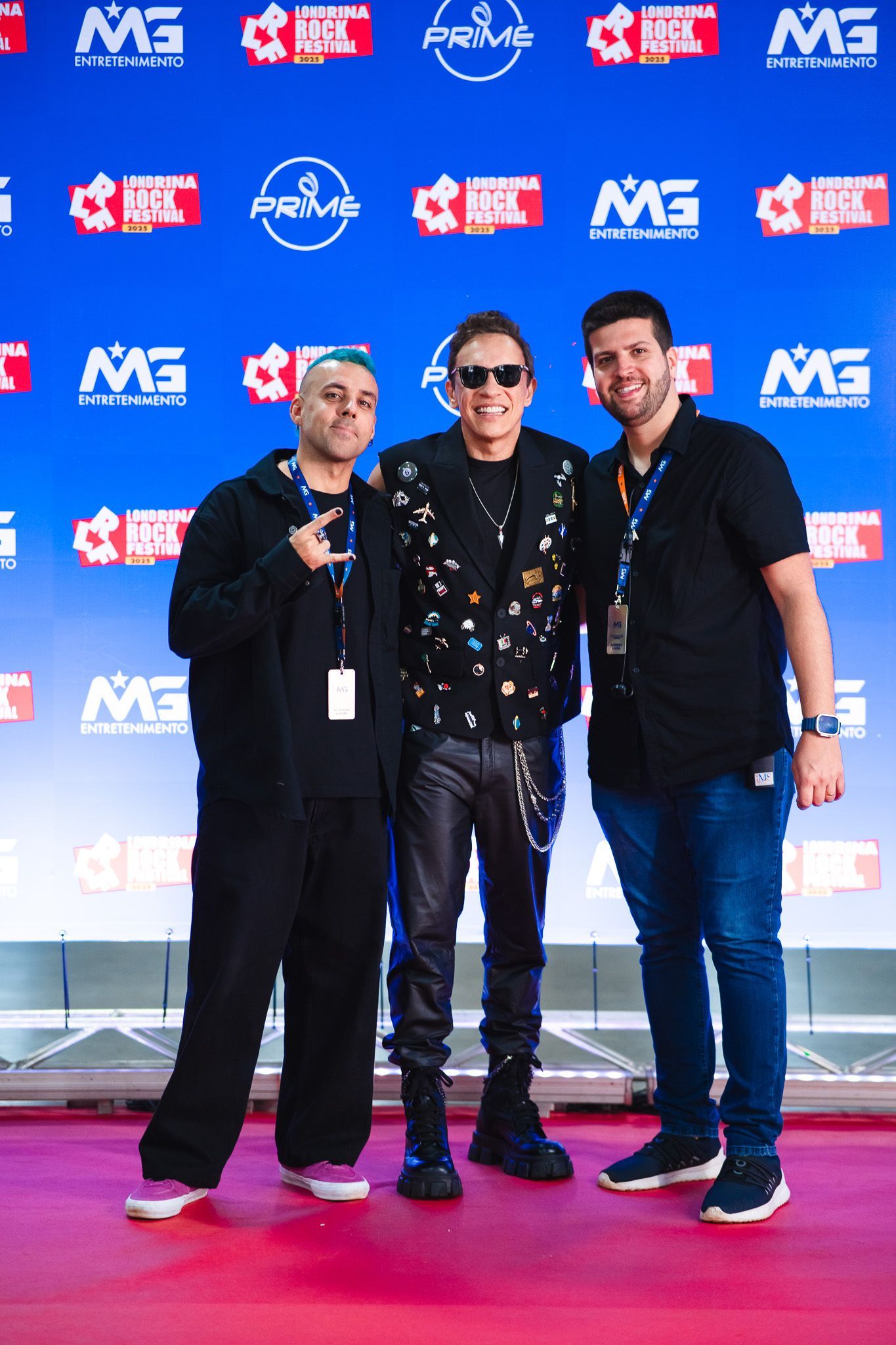 Três pessoas posando para uma foto em um tapete azul; uma vestindo calças de couro pretas, as outras com roupas casuais.