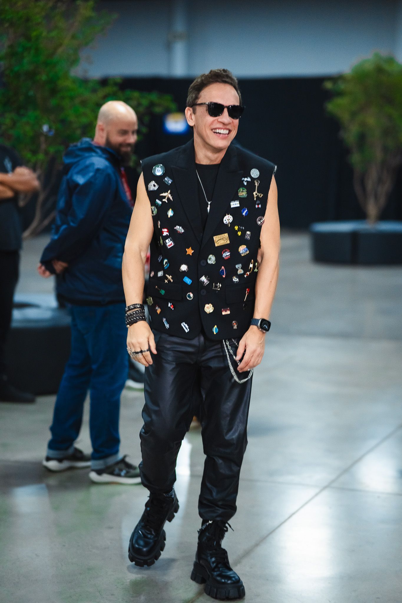 Homem de colete preto com broches, calça de couro, óculos escuros, sorrindo, caminhando em ambiente interno.