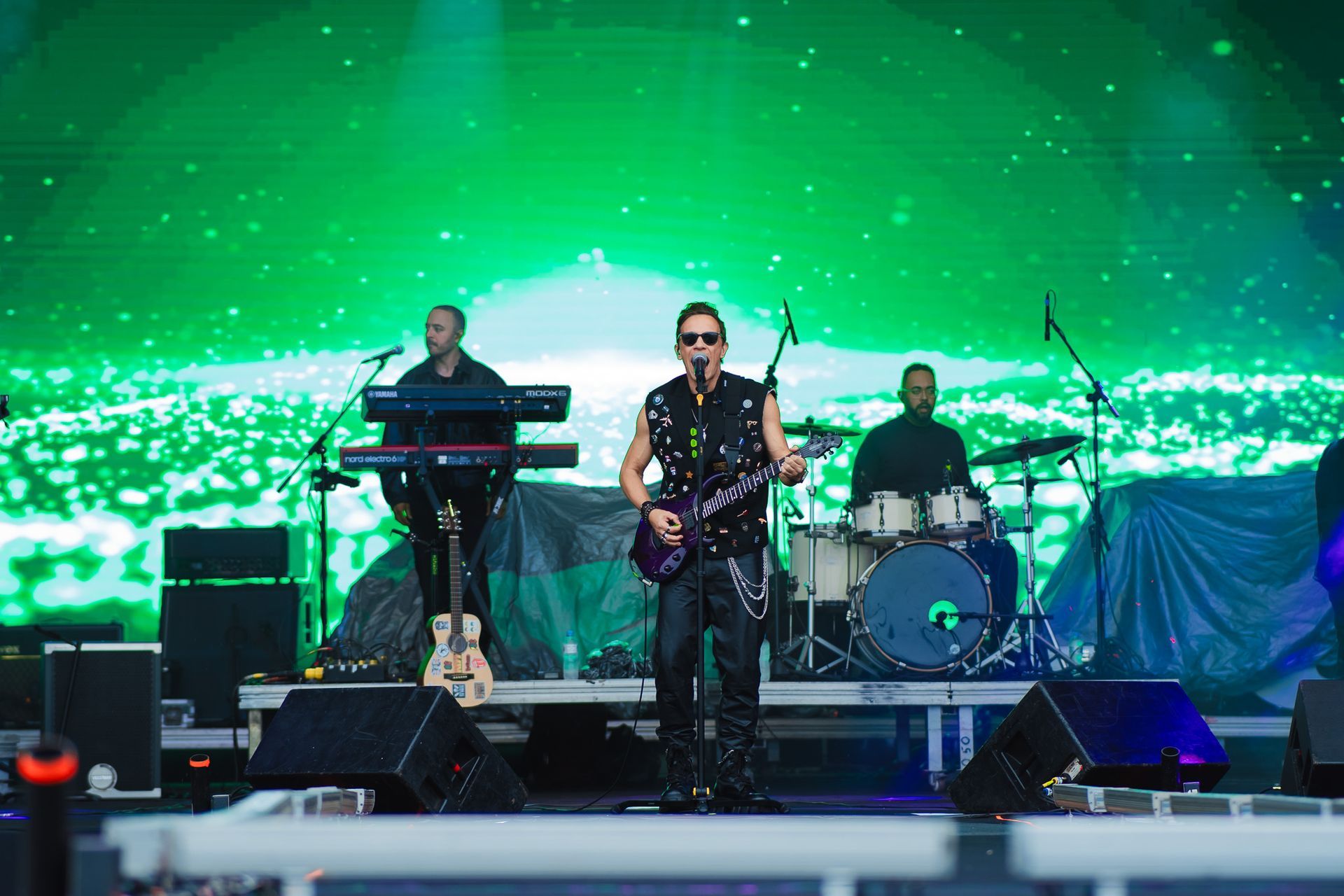 Banda se apresentando no palco com fundo verde. O vocalista toca guitarra; o tecladista e o baterista também são visíveis.