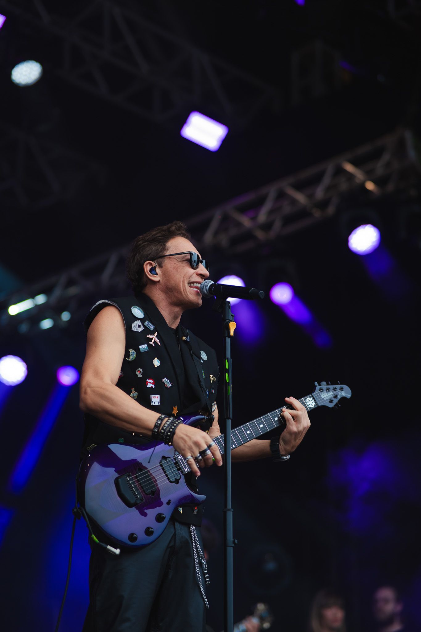 Homem tocando um baixo roxo no palco, cantando ao microfone. Ambiente escuro com luzes de palco azuis.