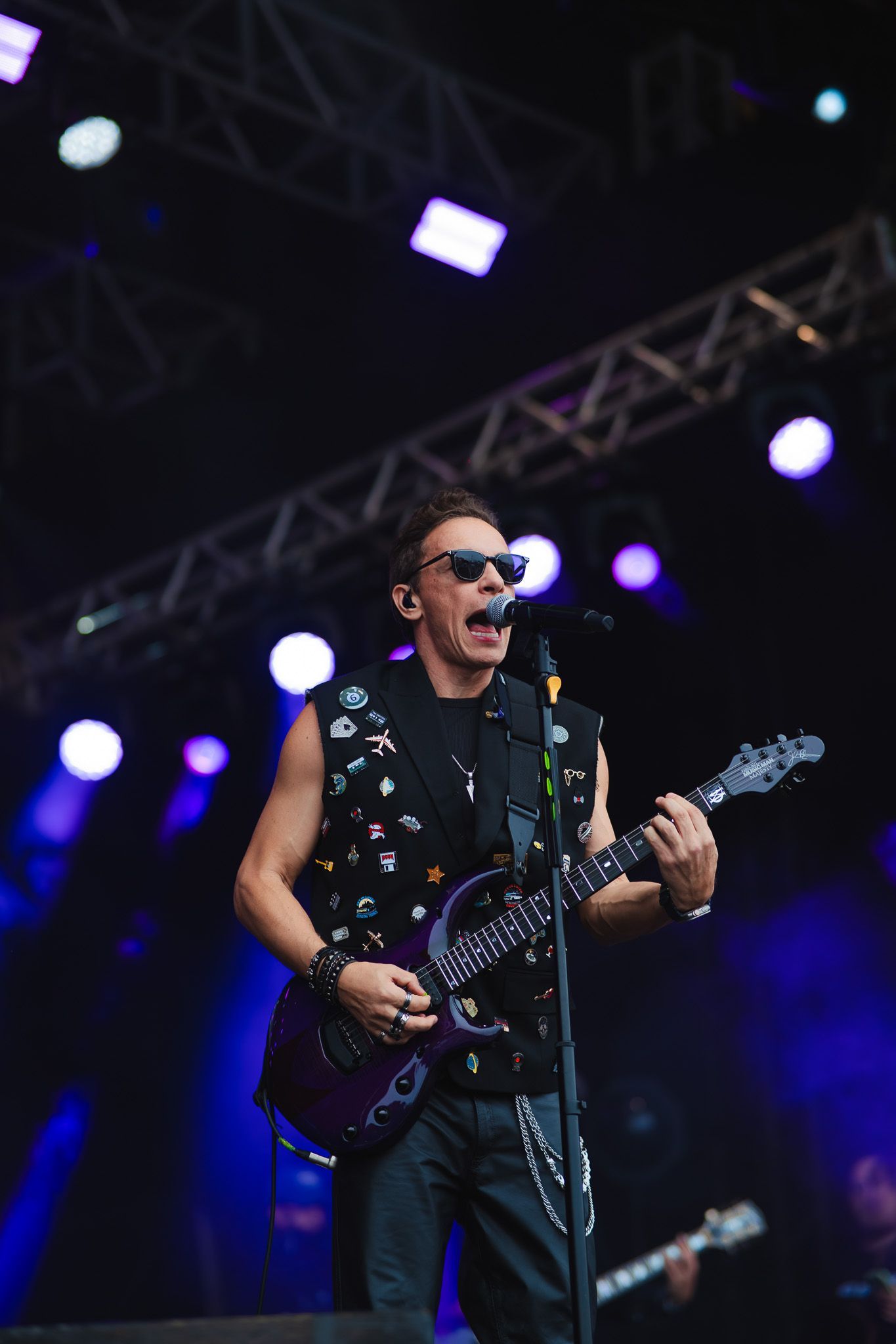 Músico no palco cantando e tocando uma guitarra roxa, usando óculos escuros e um colete com tachas, sob luzes roxas.
