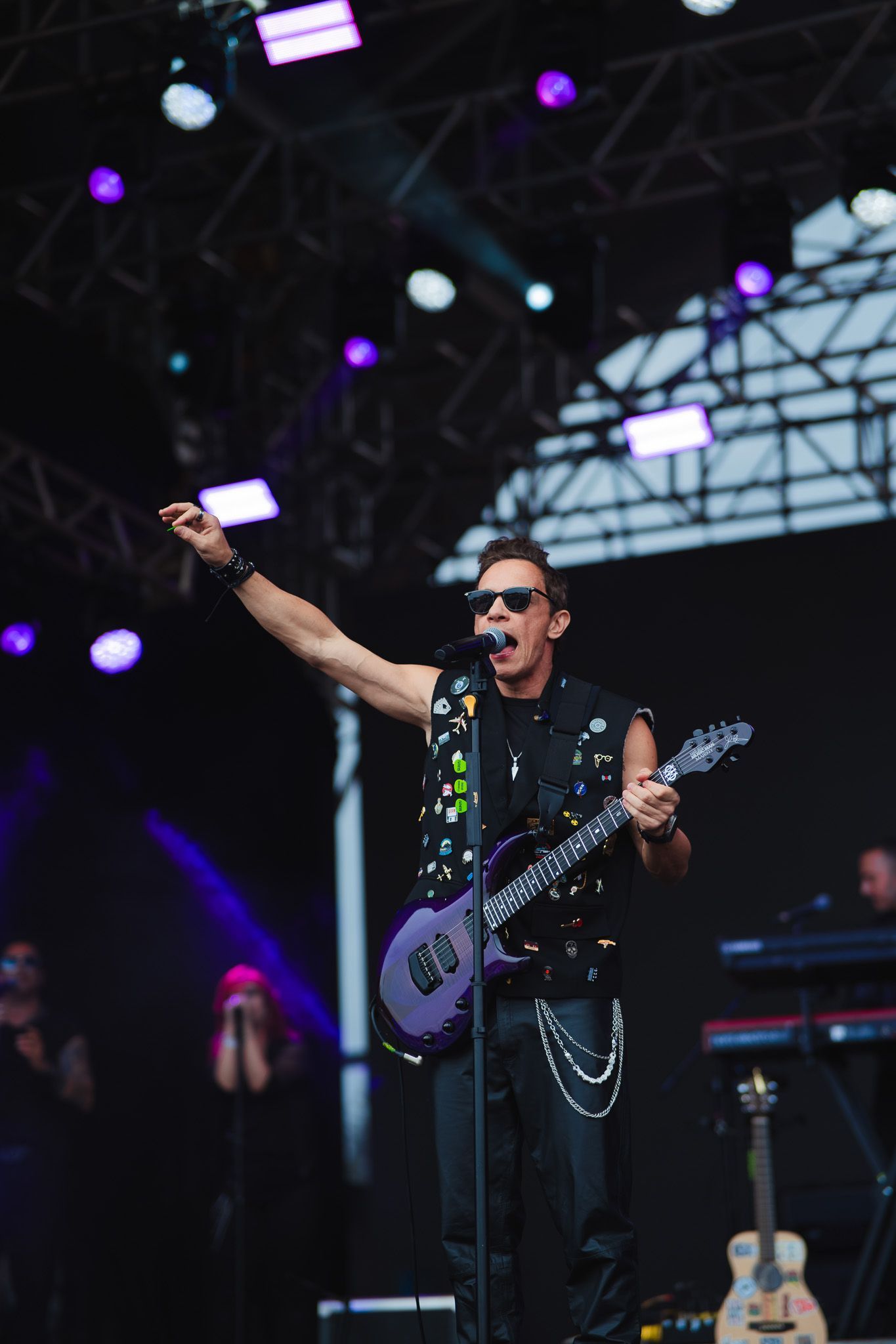 Músico no palco tocando guitarra elétrica roxa, com o braço levantado, vestindo colete e óculos escuros.