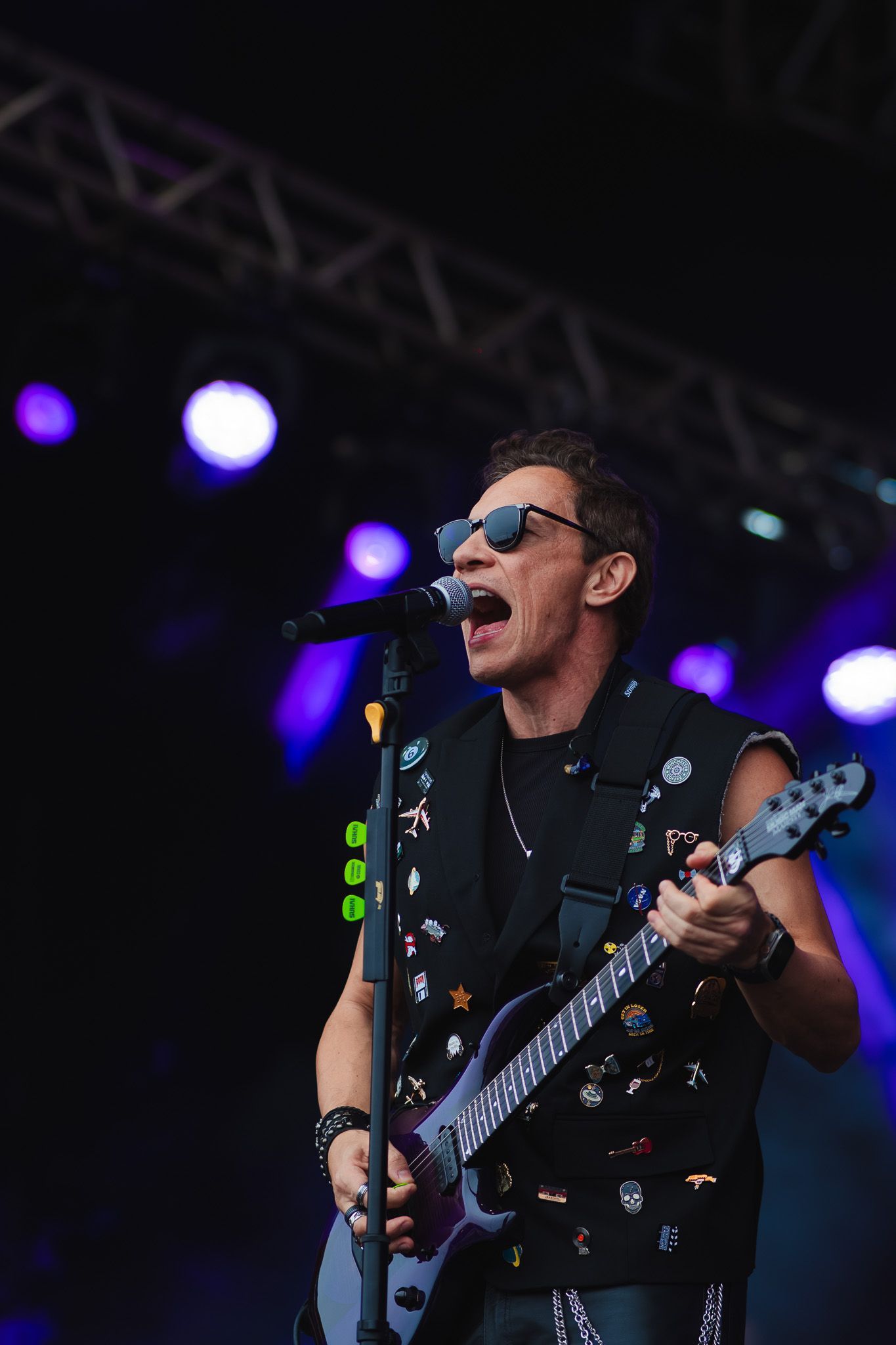 Um artista cantando e tocando guitarra elétrica no palco, usando óculos escuros e um colete com tachas, sob luzes de palco roxas.