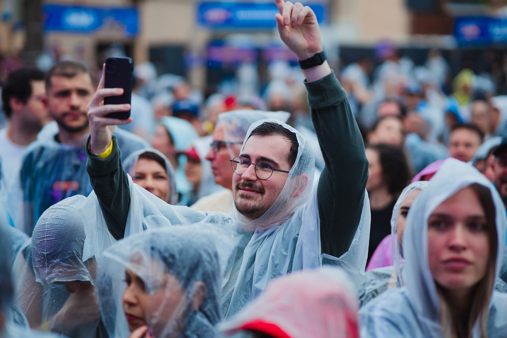 Um homem na multidão segura o celular, vestindo uma capa de chuva e sorrindo. Outras pessoas com capas de chuva o cercam.