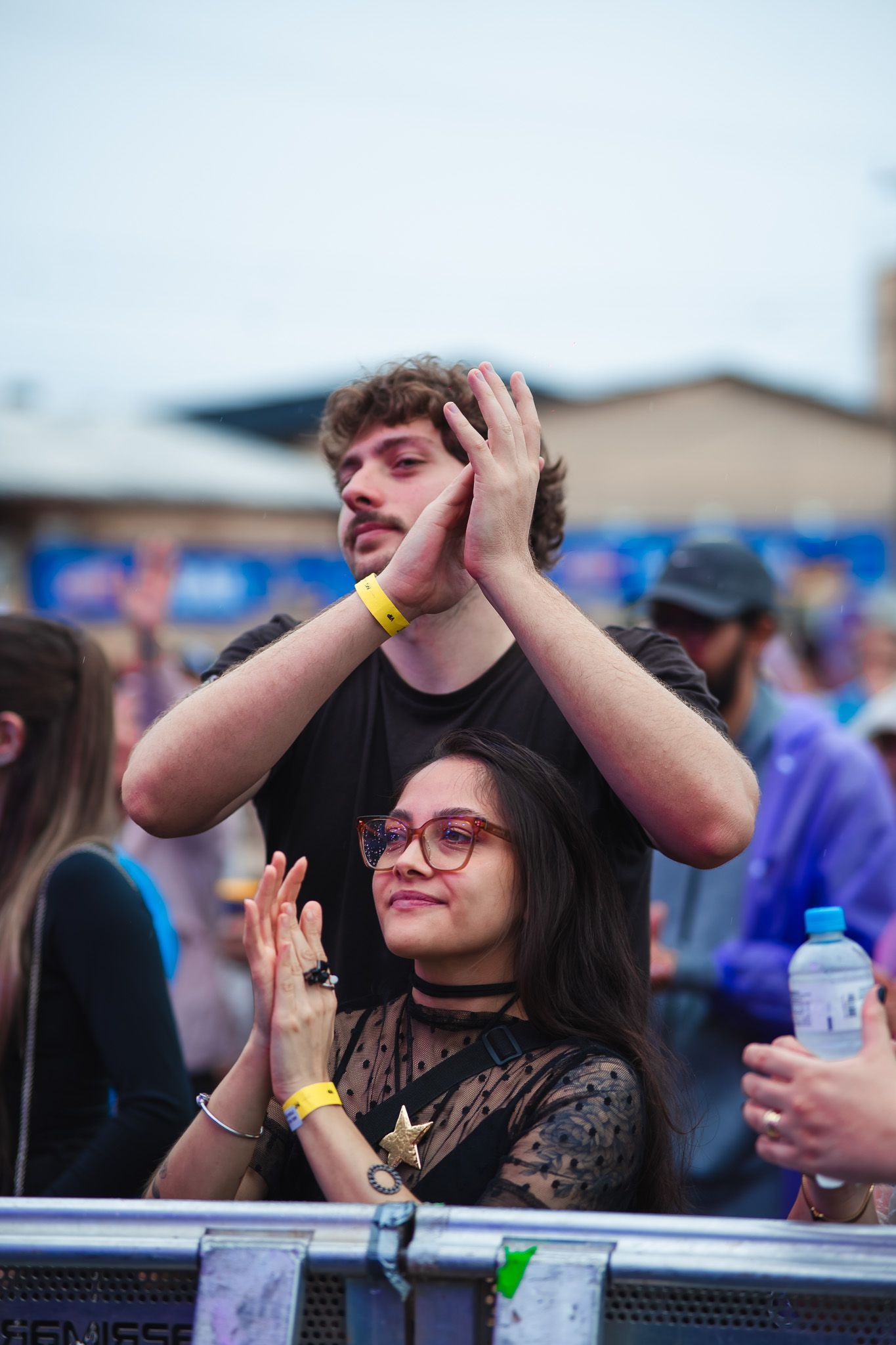 Duas pessoas aplaudem em um evento ao ar livre. O homem tem cabelo castanho e camisa preta. A mulher de óculos preta sorri.