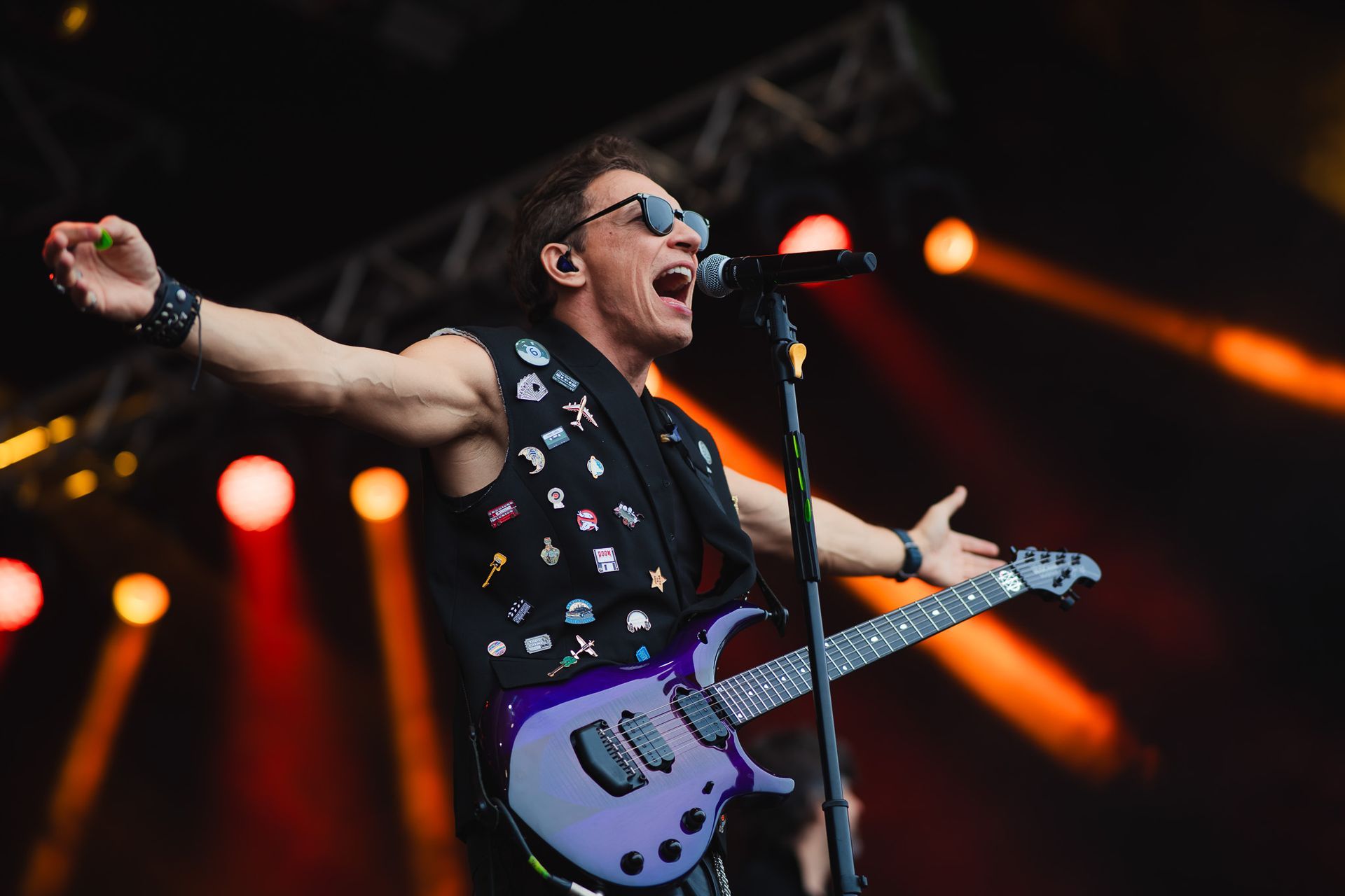 Um artista cantando e tocando guitarra elétrica no palco, com os braços estendidos, sob luzes de palco.