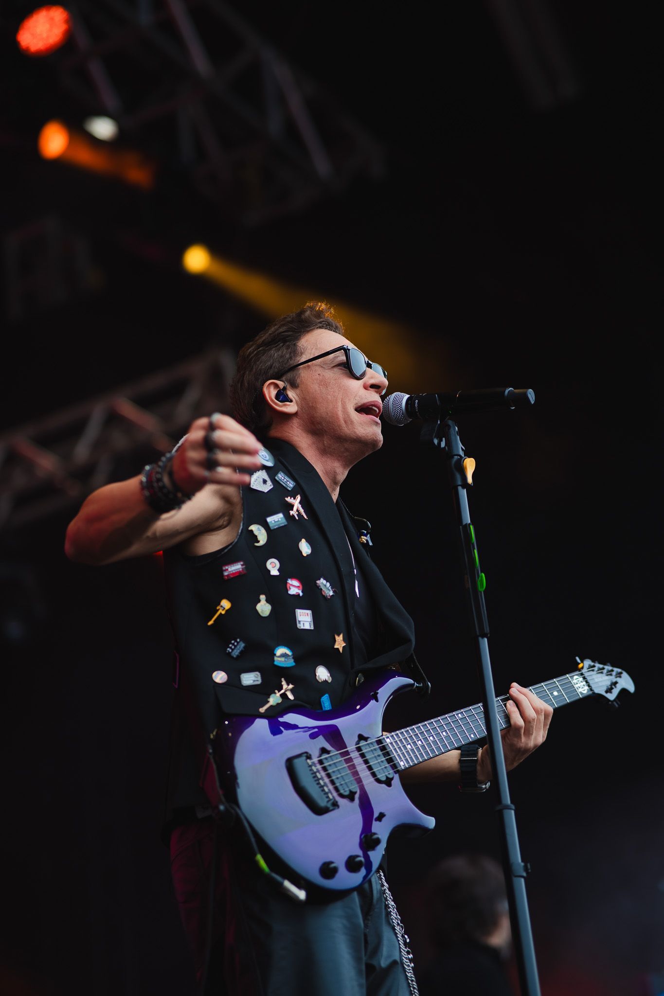 Um artista tocando guitarra roxa no palco, vestindo um colete coberto de broches e óculos escuros, cantando em um microfone.