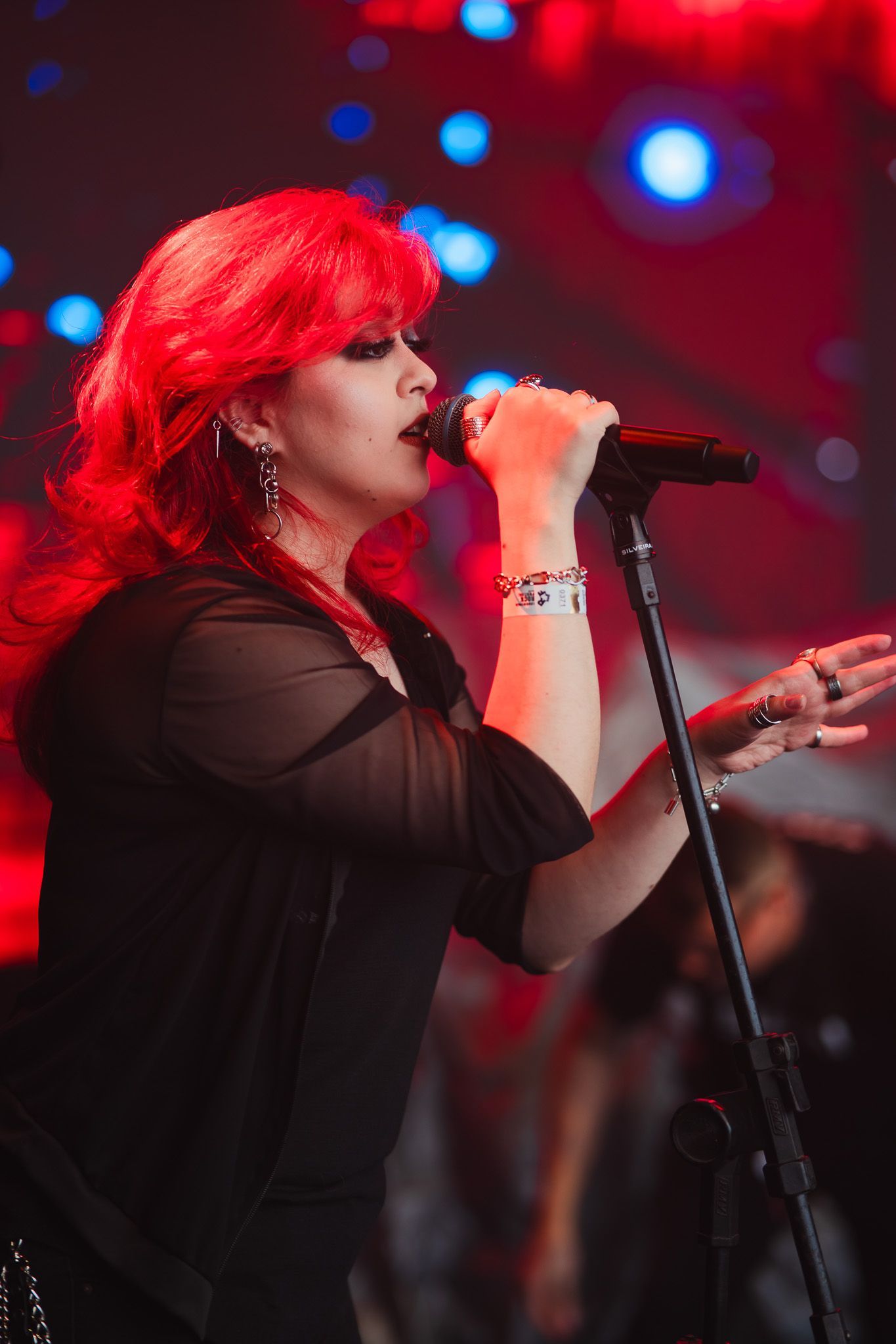 Mulher ruiva cantando ao microfone no palco, iluminada por luzes vermelhas e azuis.