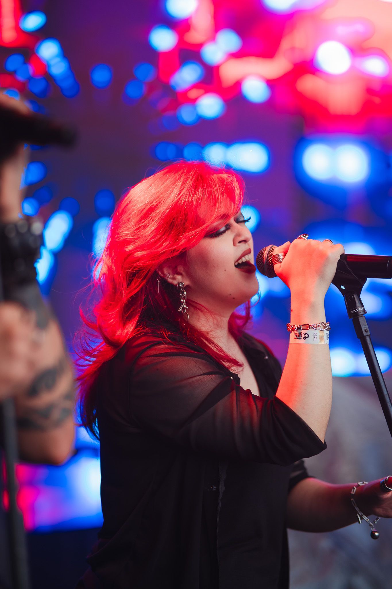 Mulher de cabelos ruivos vibrantes cantando ao microfone no palco; luzes azuis e vermelhas ao fundo.