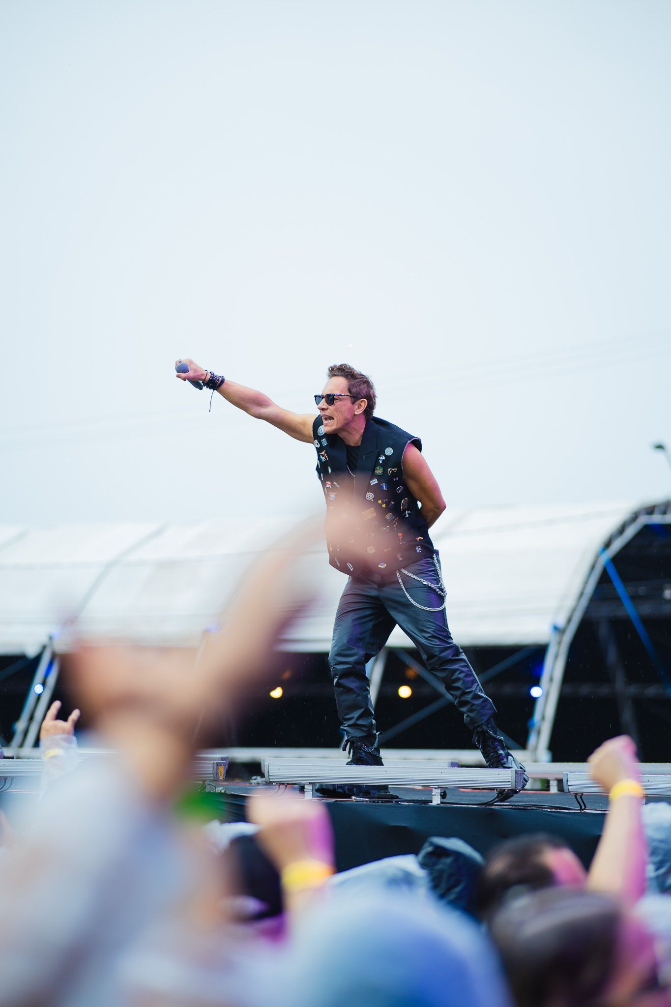 Um artista no palco, de colete, óculos escuros e punho erguido, com a multidão em primeiro plano. Ambiente externo, céu nublado.