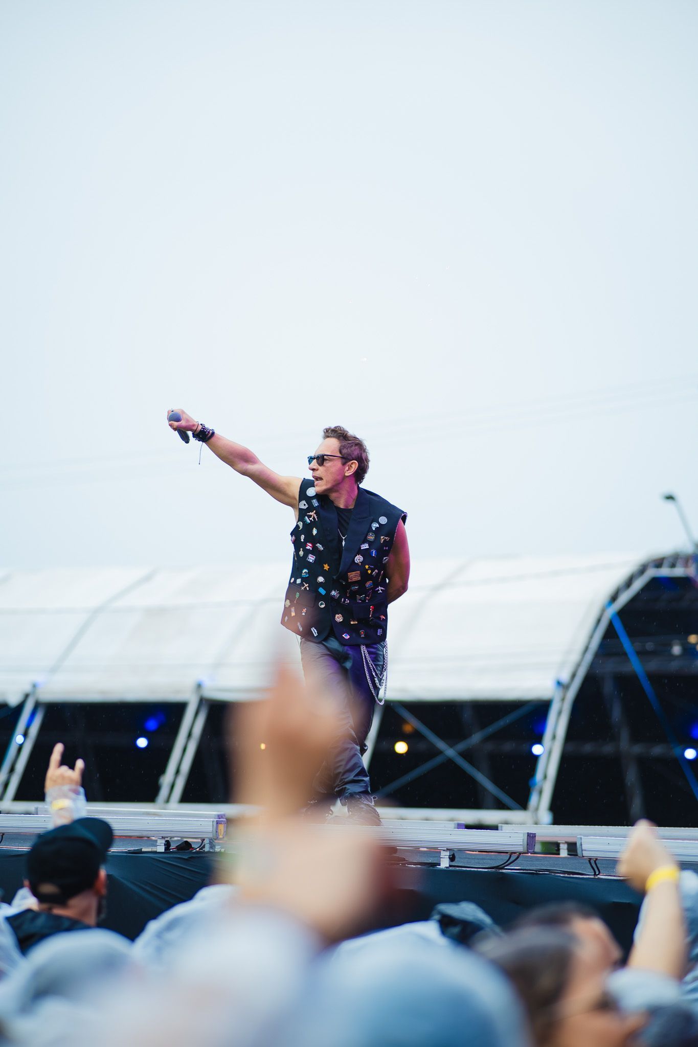 Cantor se apresentando no palco, usando blusa regata estampada e óculos de sol, braço levantado, plateia em primeiro plano.