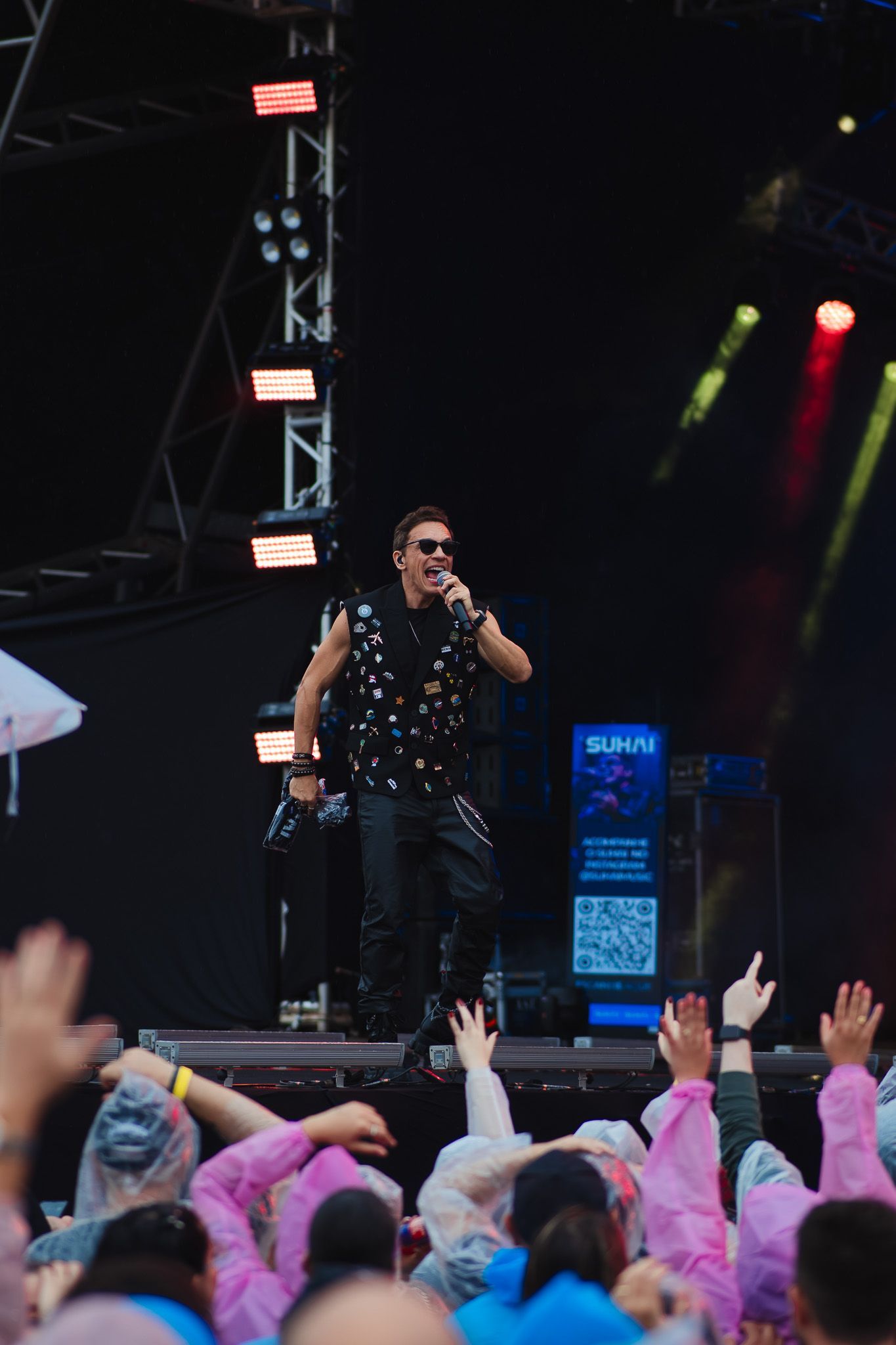 Um artista cantando no palco, usando óculos escuros e blusa com tachas, plateia com as mãos erguidas, iluminação de palco.