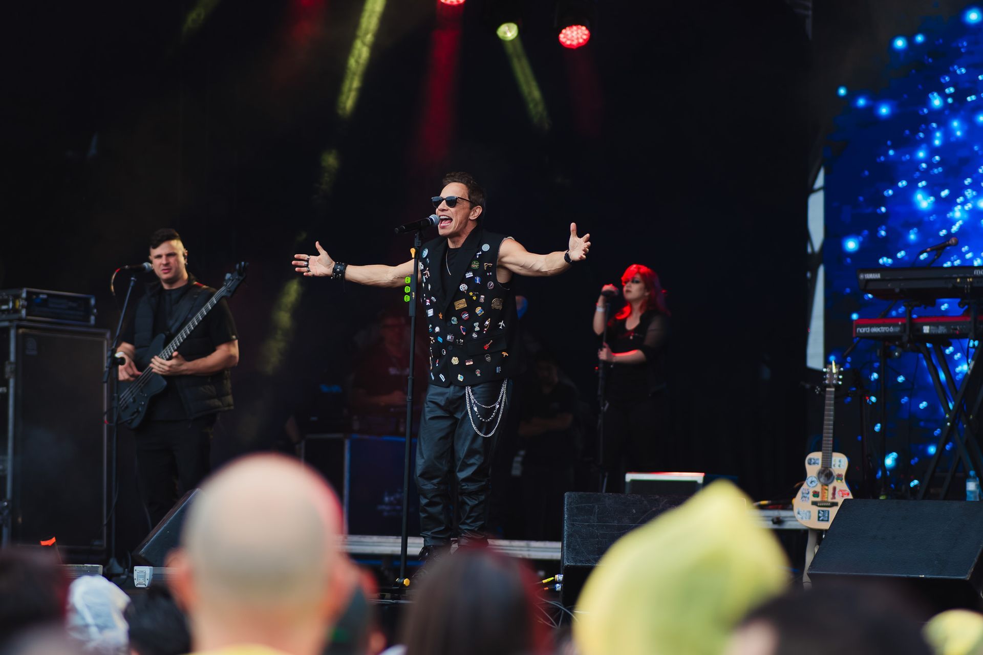 Banda se apresentando no palco em um show ao ar livre. Vocalista com colete e calça de couro com os braços estendidos. 