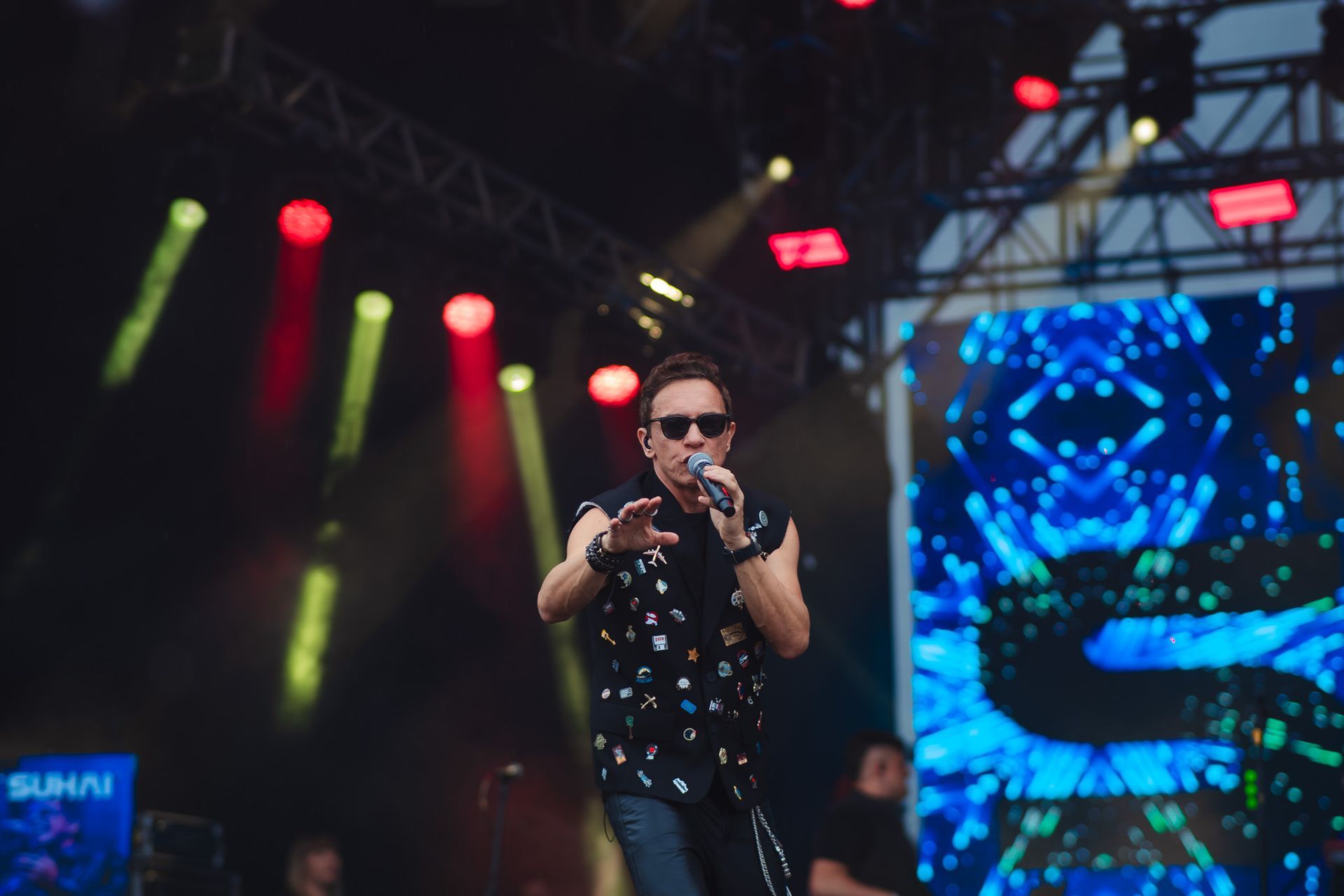 Um artista cantando no palco, usando óculos escuros e blusa sem mangas, com luzes de palco e um telão ao fundo.