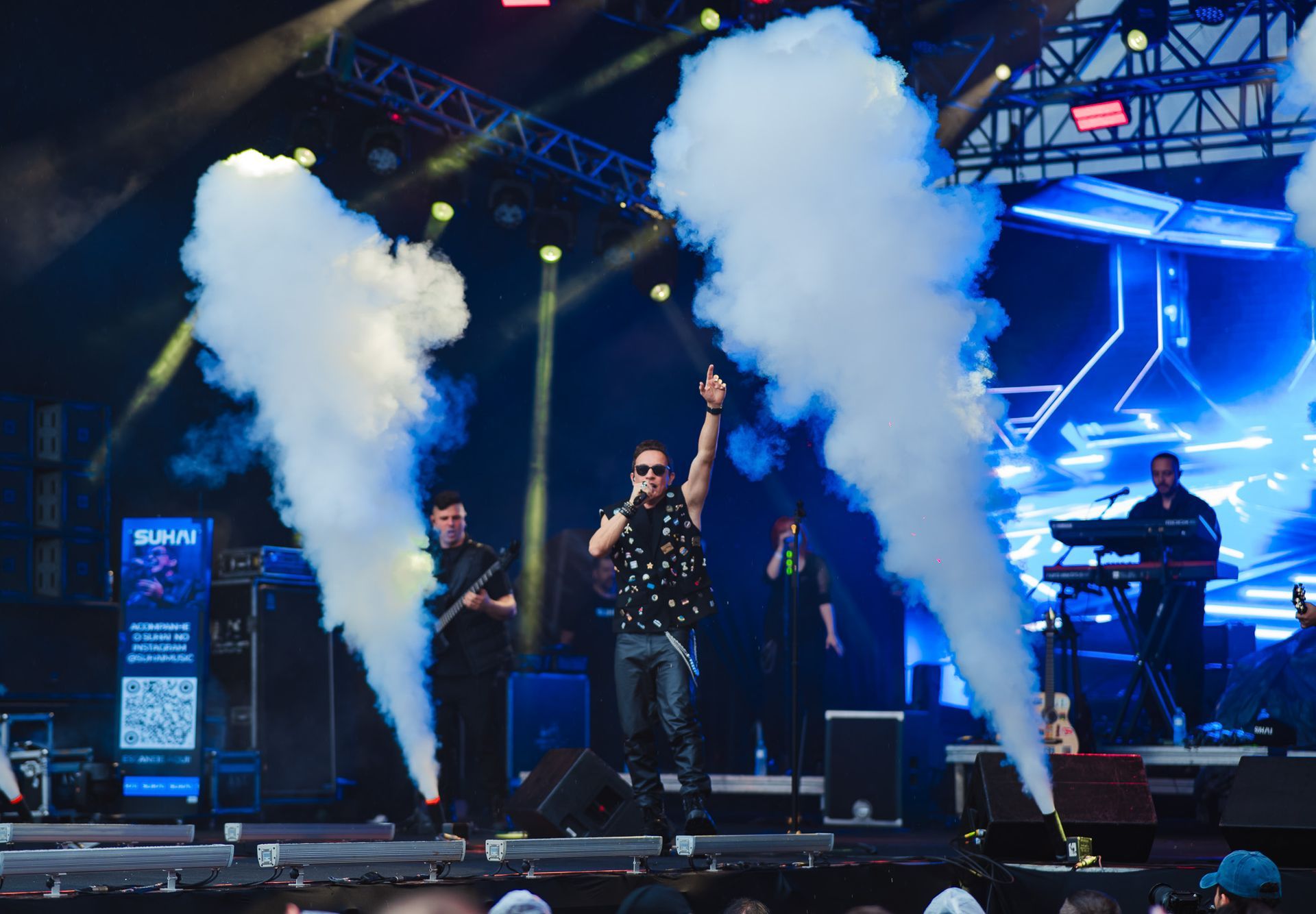Banda se apresentando no palco. O vocalista de óculos escuros levanta o braço, acompanhado por outros integrantes da banda.