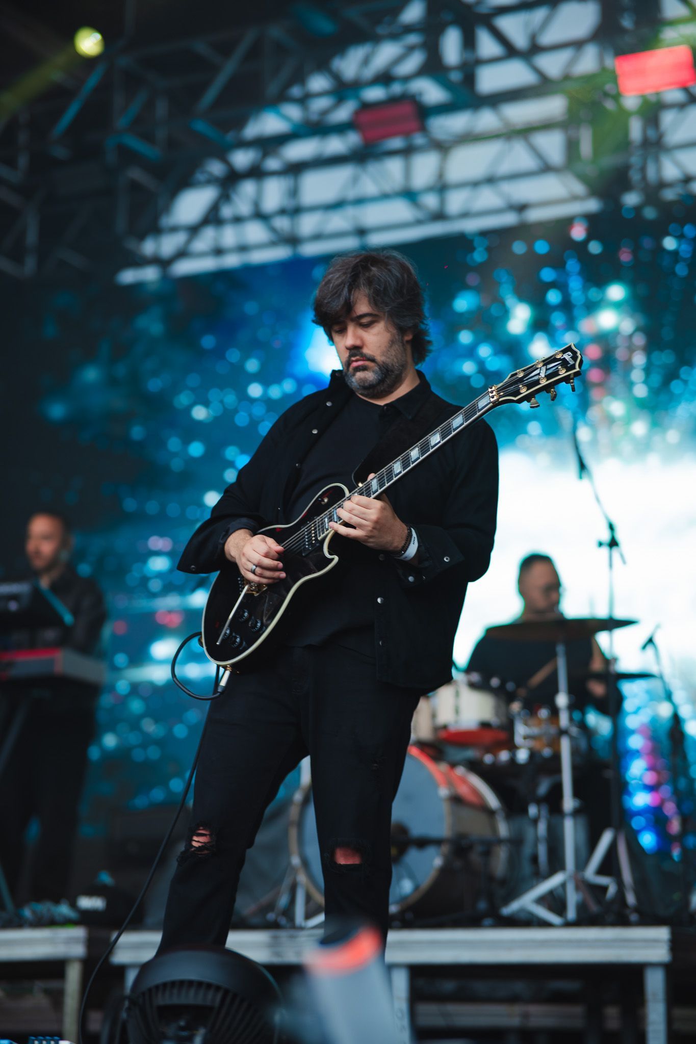 Guitarrista vestido de preto tocando no palco; banda ao fundo com bateria e teclado.
