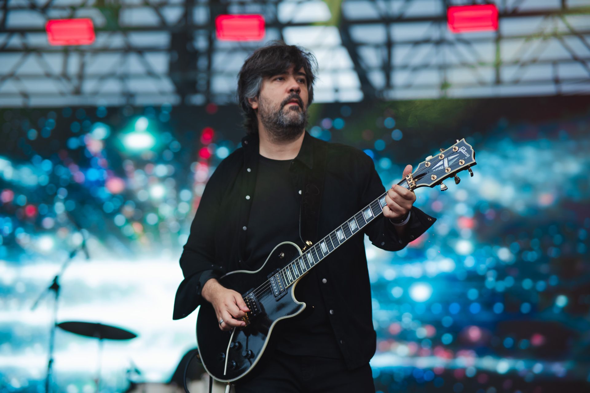 Guitarrista tocando uma guitarra preta no palco, vestindo uma camisa preta, com um cenário colorido ao fundo.