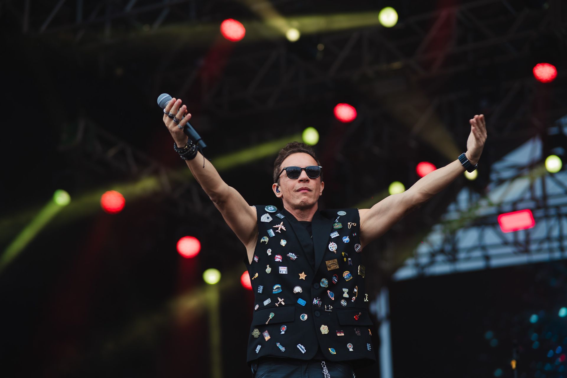Um artista no palco com os braços levantados, usando óculos escuros, colete coberto de pins, microfone na mão e luzes de palco