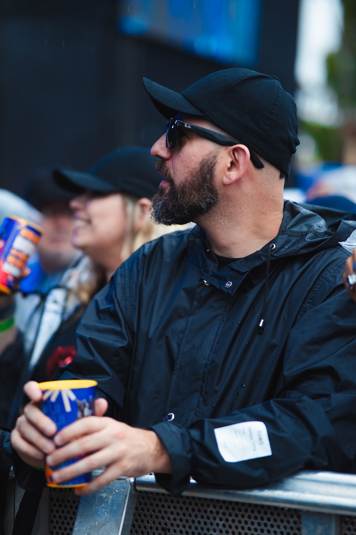 Homem de chapéu e jaqueta pretos, segurando uma caneca, olhando para o lado, para fora, em direção a uma cerca.