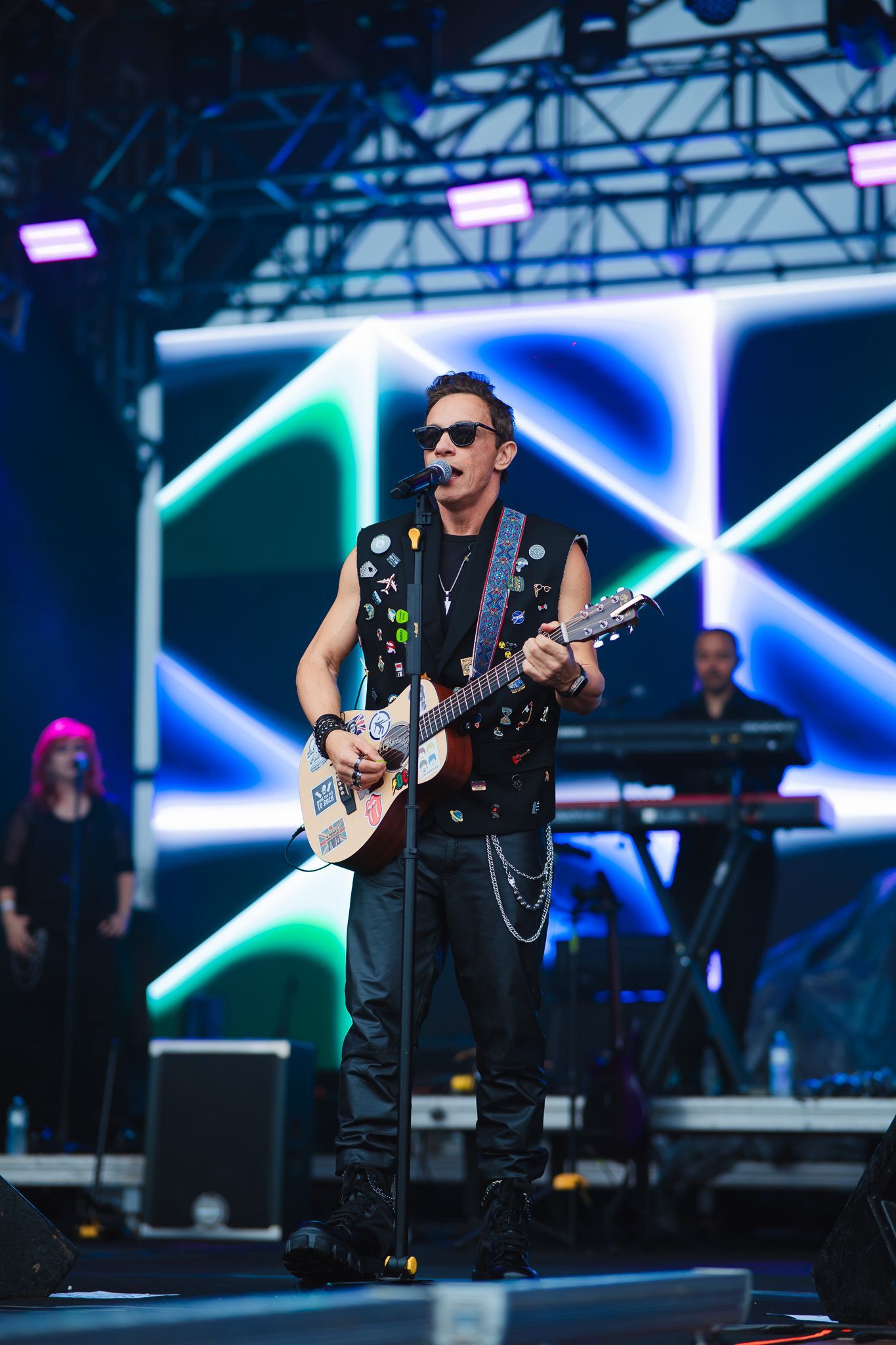 Um artista tocando guitarra no palco, vestindo um colete com tachas e óculos escuros, com um fundo azul e verde.