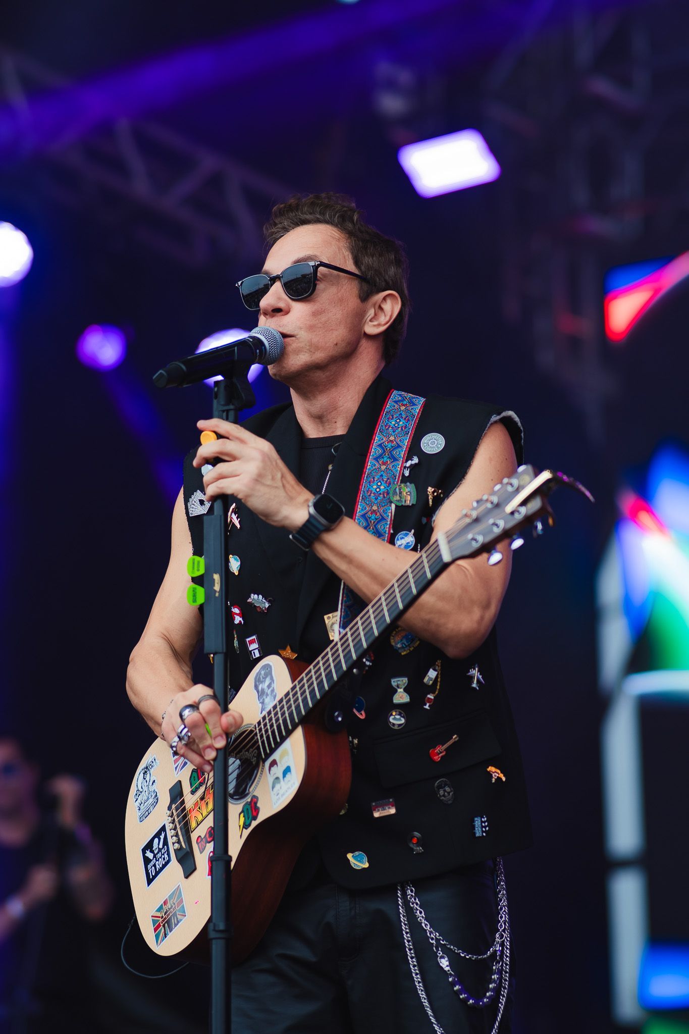 Músico no palco cantando e tocando violão, usando óculos escuros e um colete adornado com broches, sob luzes azuis de palco.