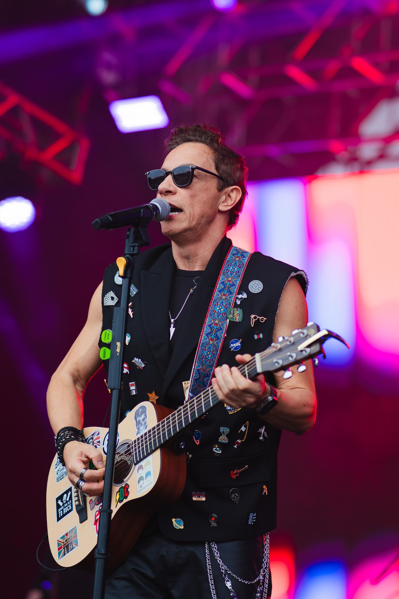 Um artista no palco cantando e tocando violão. Usando óculos escuros e colete coberto de pins.