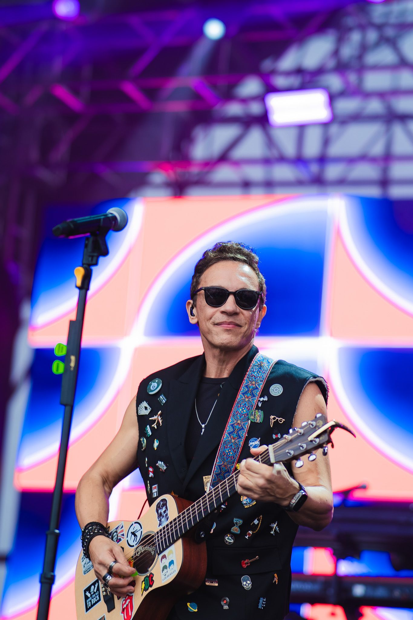 Um artista de óculos escuros tocando guitarra no palco, vestindo um colete preto, com um cenário colorido ao fundo.