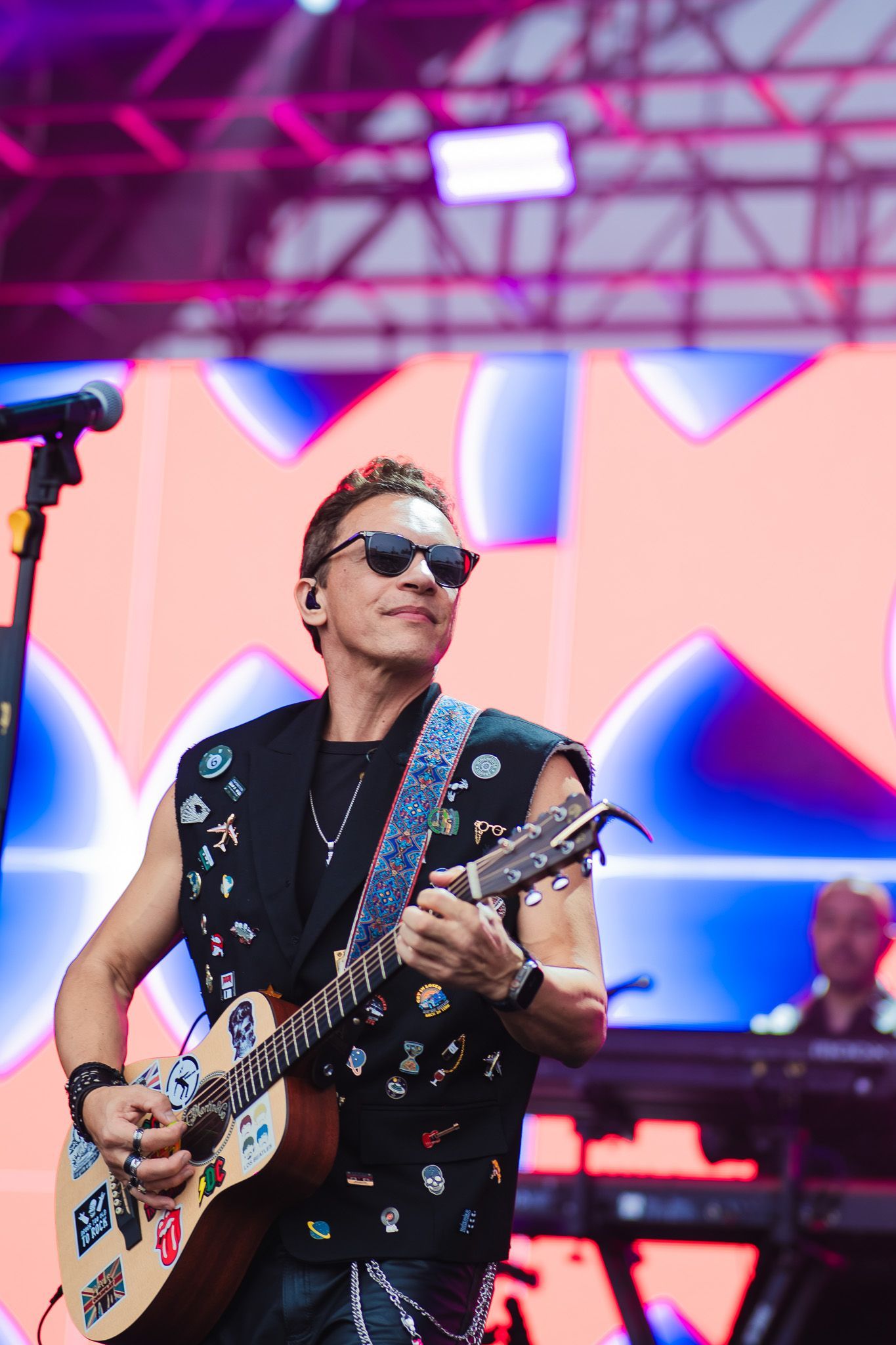 Um artista tocando guitarra no palco, usando óculos escuros e um colete com tachas. Fundo colorido.