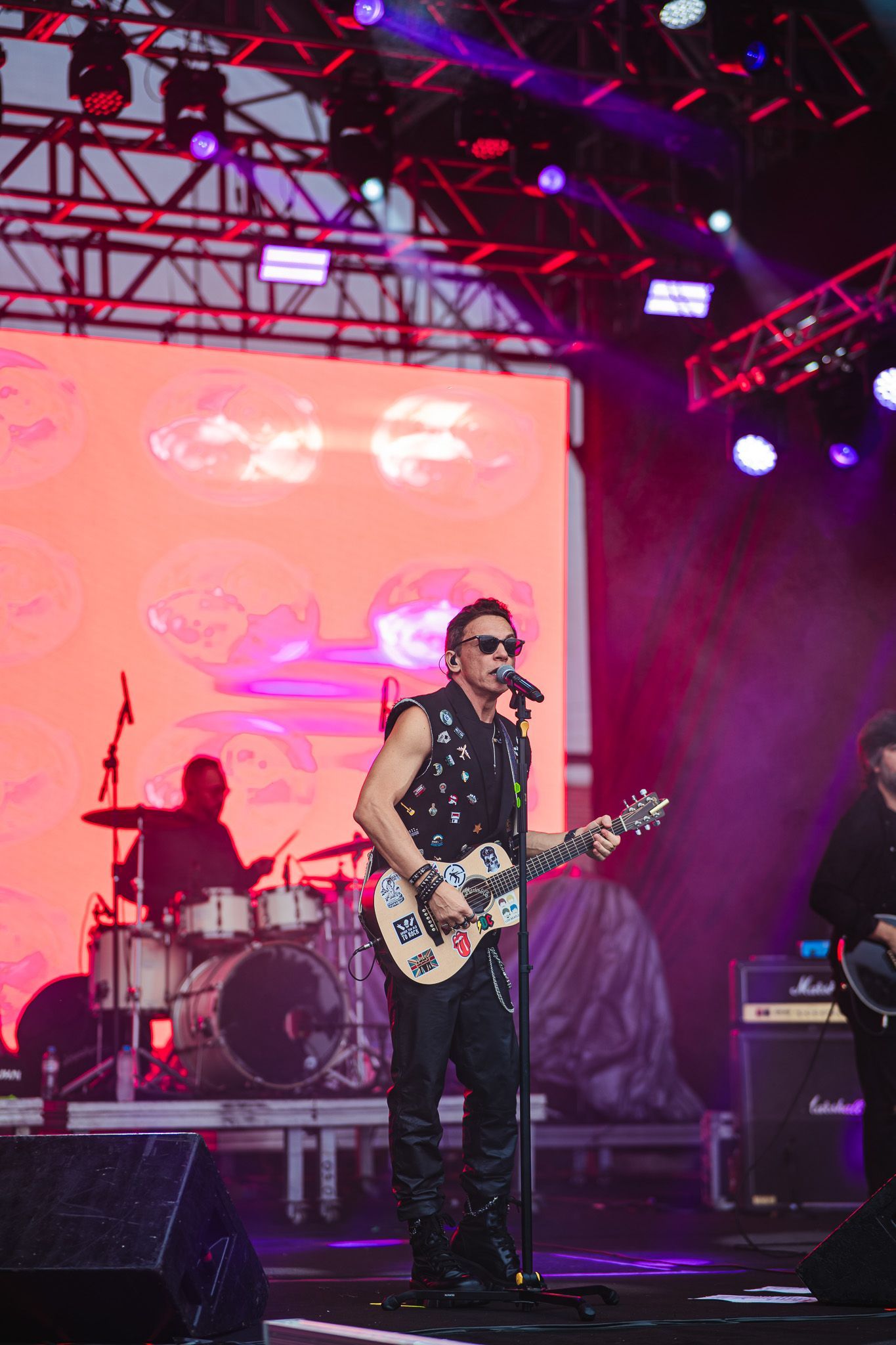 Uma banda se apresenta no palco. Um homem de óculos escuros toca guitarra e canta, com um baterista e outros membros da banda