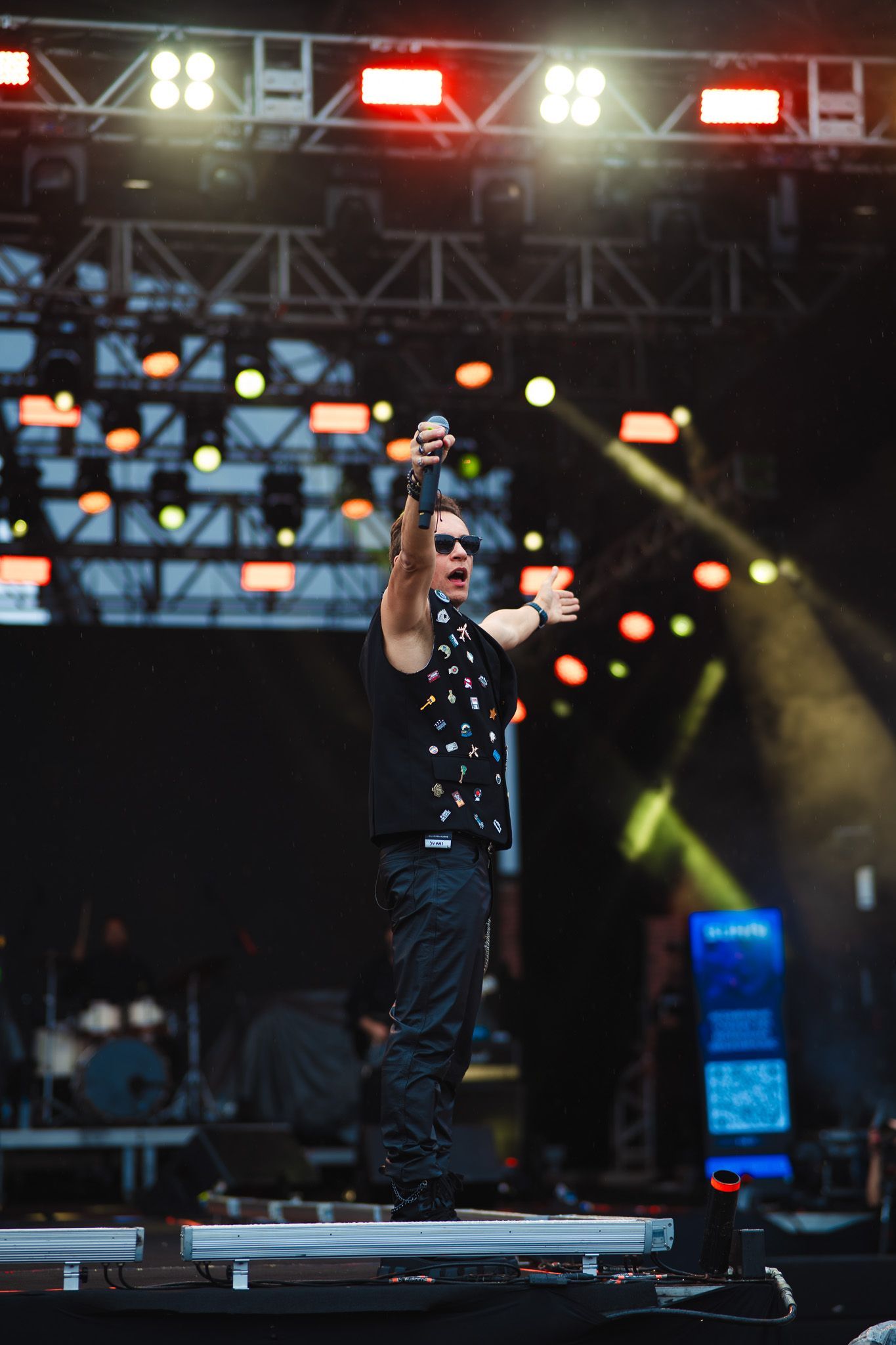 Um artista no palco, com os braços erguidos, vestindo colete preto com tachas. Luzes de palco ao fundo.