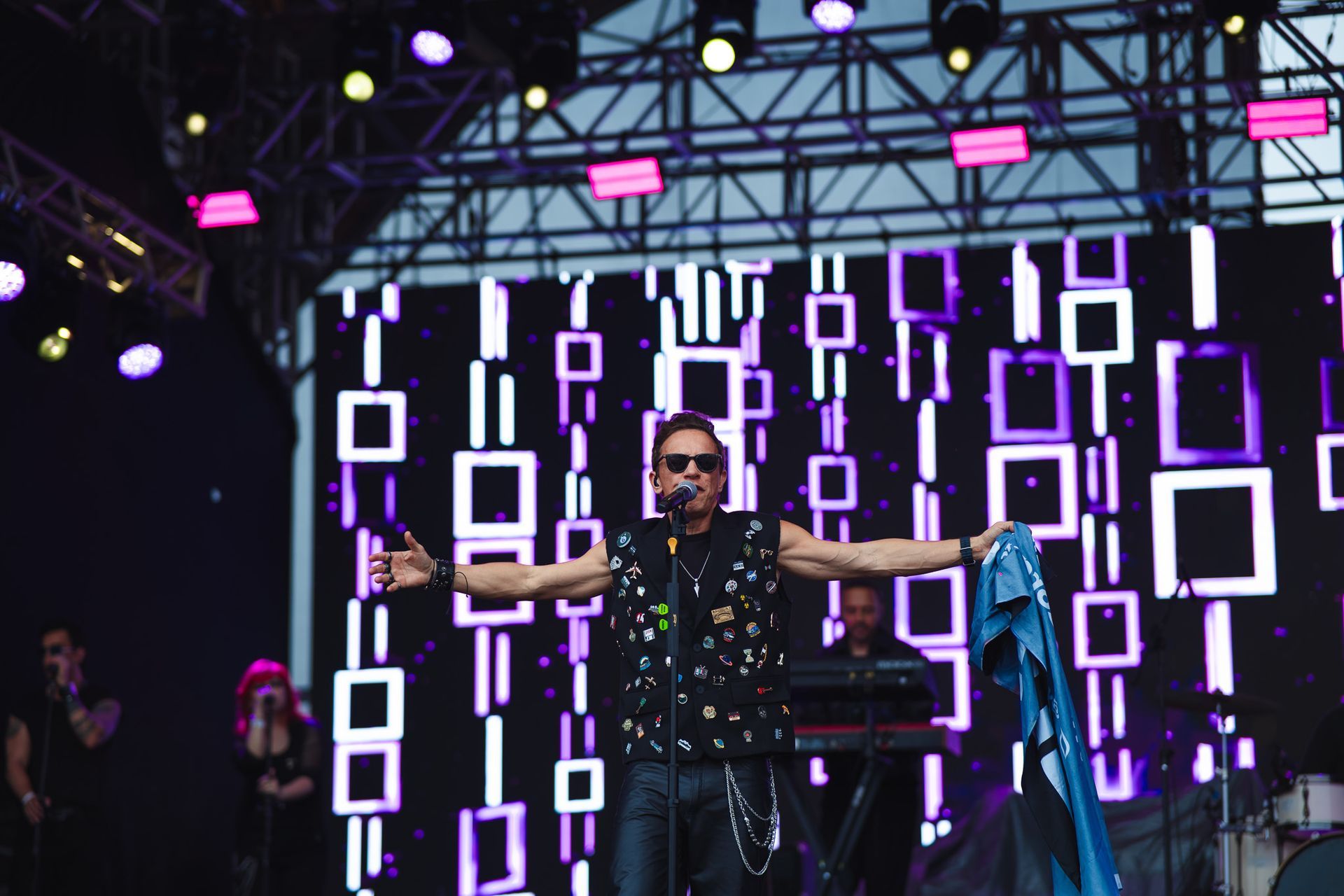 Um artista no palco com os braços estendidos, usando óculos escuros e colete preto. Cenário de palco com luzes roxas e brancas.