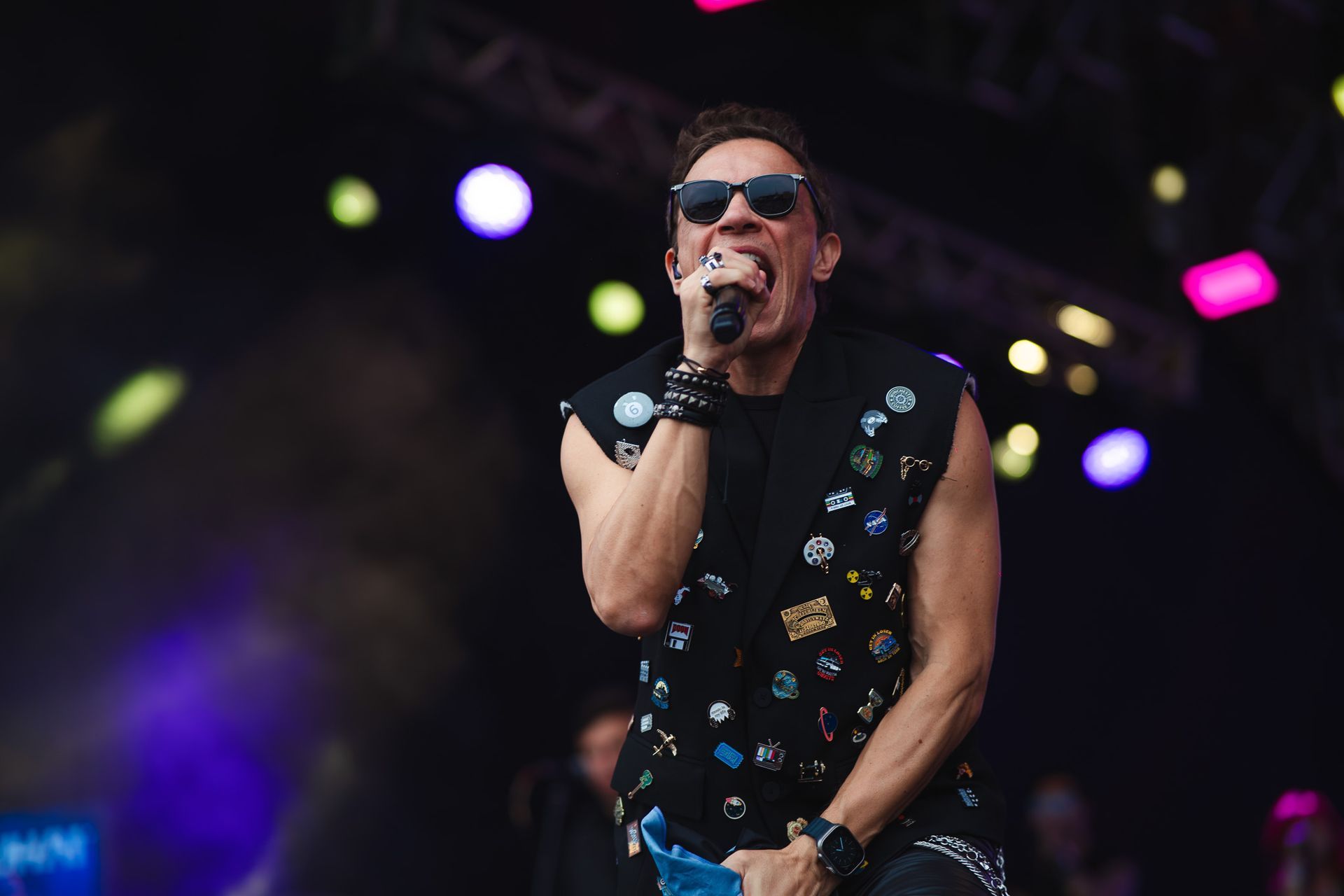 Um artista cantando em um microfone no palco, usando óculos escuros e colete preto com broches. Luzes de palco roxas e rosas.