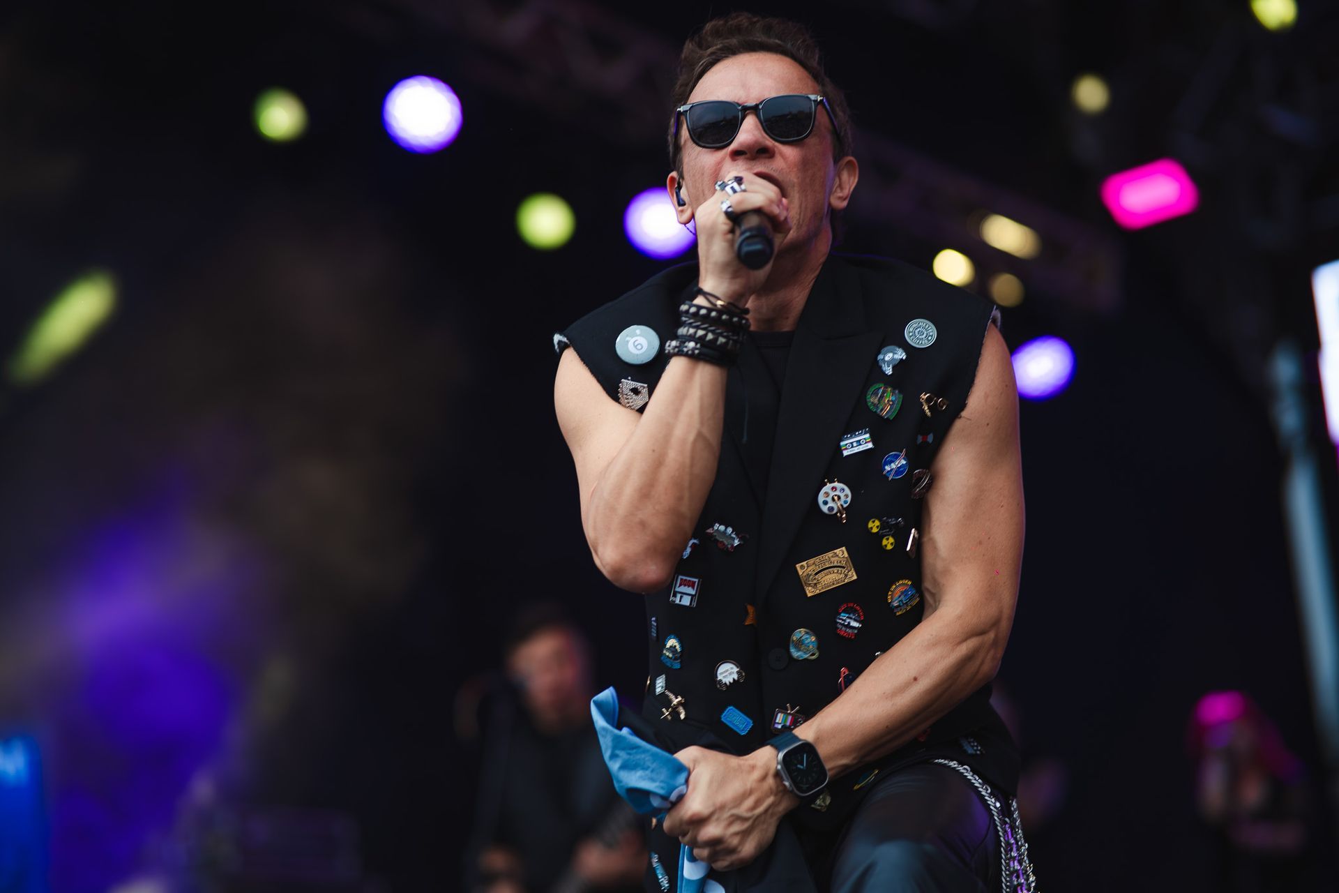 Um artista cantando em um microfone no palco, vestindo um colete preto com broches e óculos escuros.