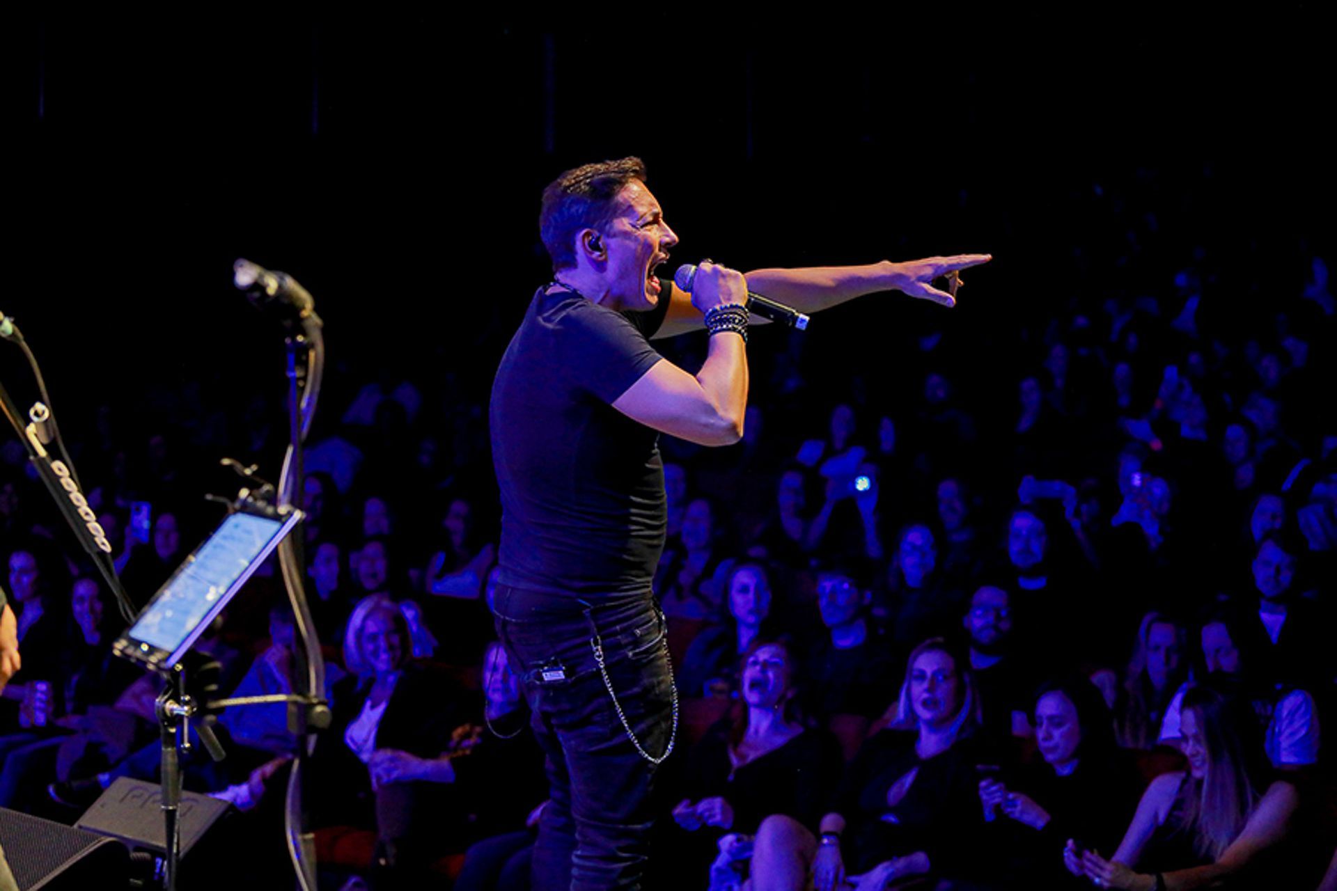 Um cantor de camisa preta segura um microfone e aponta para a plateia durante uma apresentação em um palco com pouca luz.