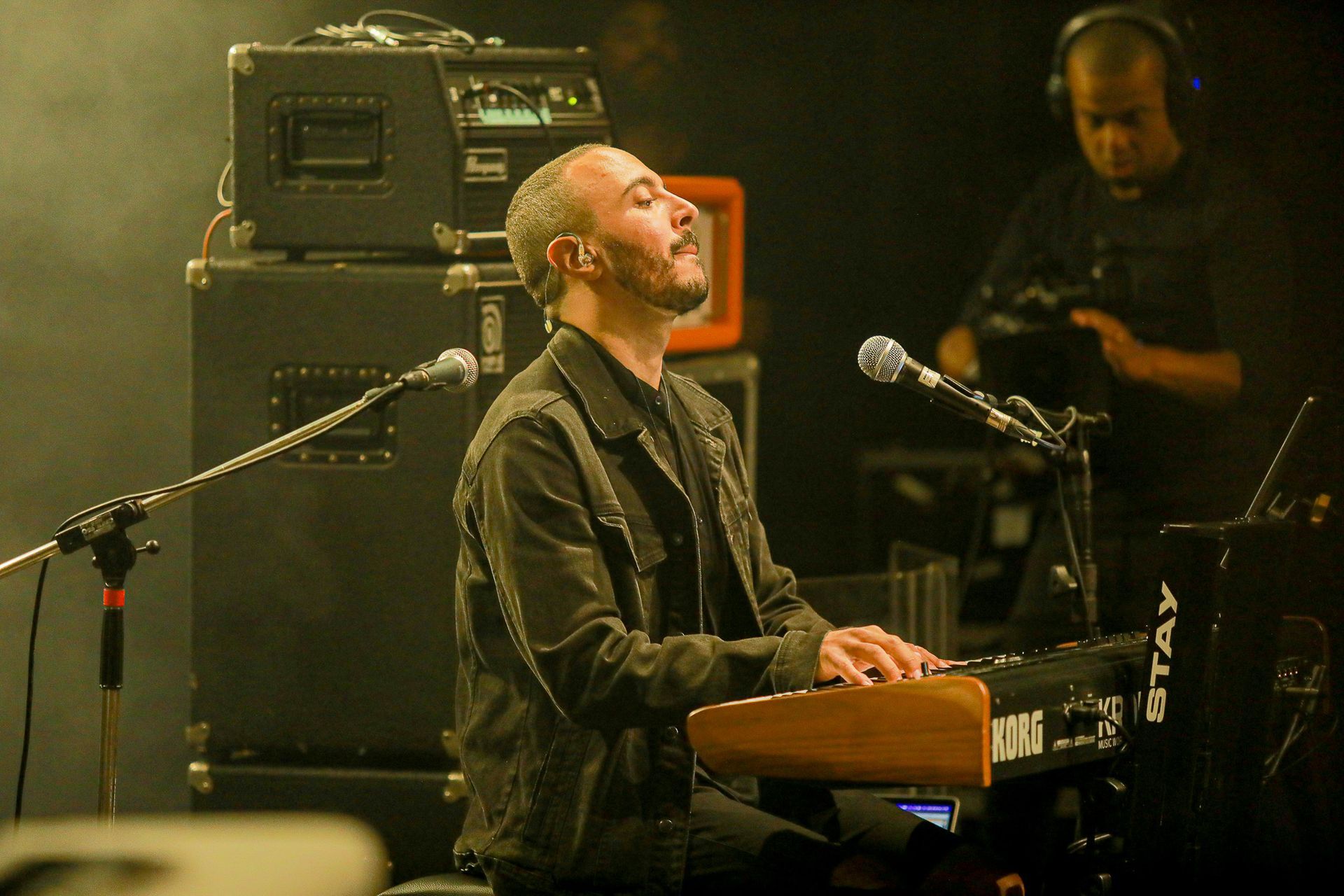 Um artista tocando teclado e cantando em um microfone em um palco com equipamento de som ao fundo.