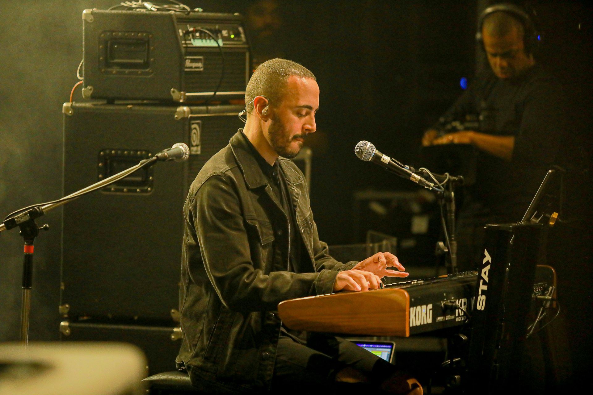 Um artista tocando teclado em um microfone em um palco com equipamento de som ao fundo, em um ambiente pouco iluminado.