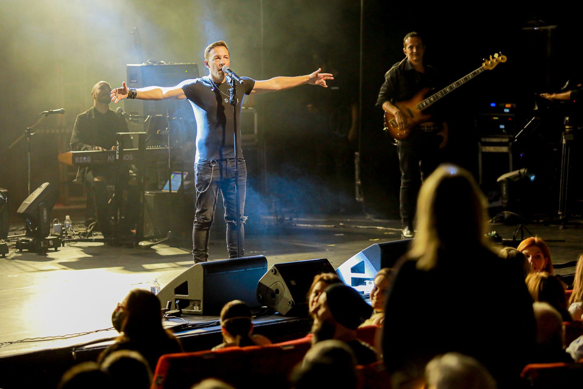 Cantor no palco, braços estendidos, apresentando-se com a banda, iluminada por luzes de palco. Público em primeiro plano.