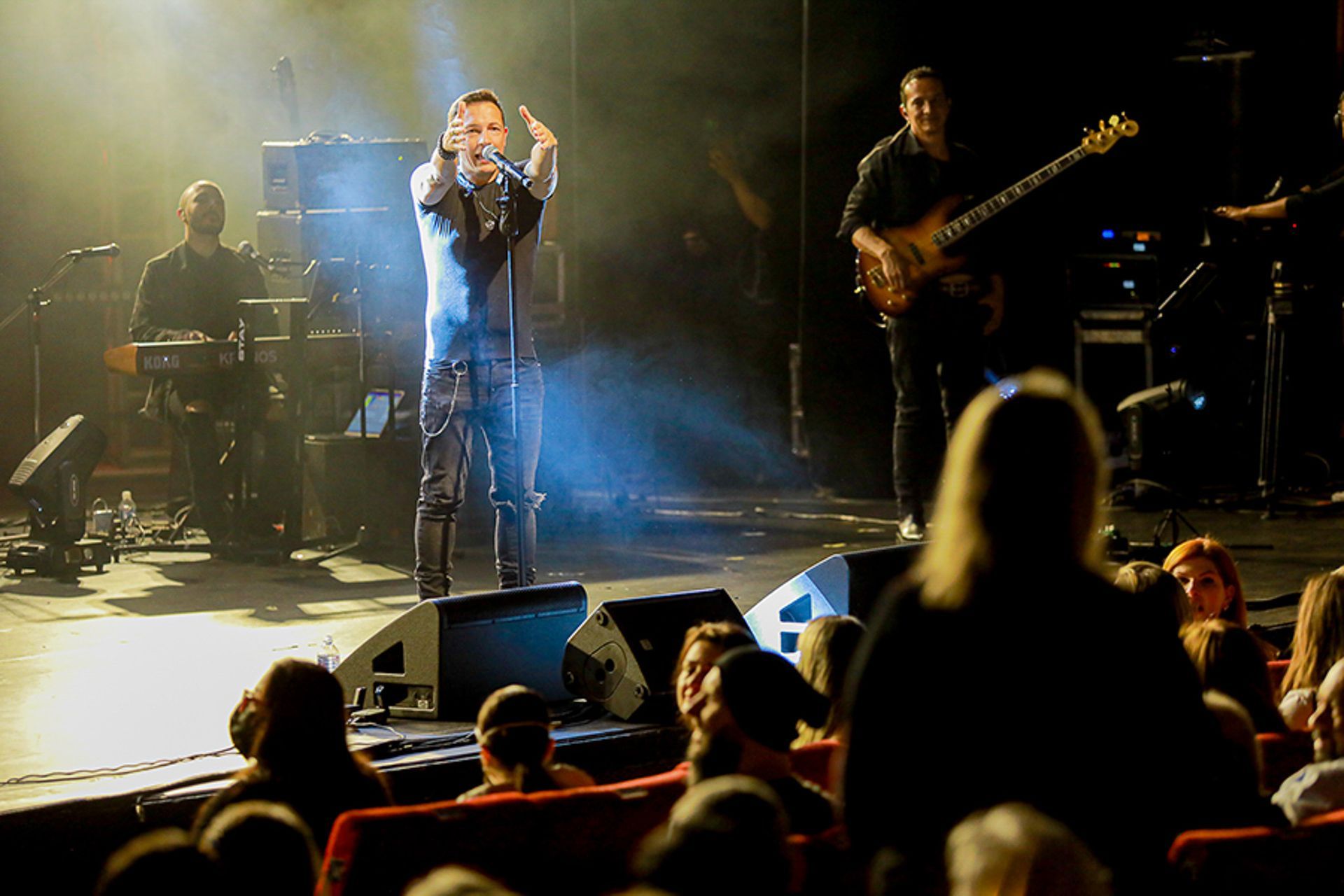 O vocalista principal se apresenta no palco com um baixista e um tecladista, vistos da plateia em um teatro
