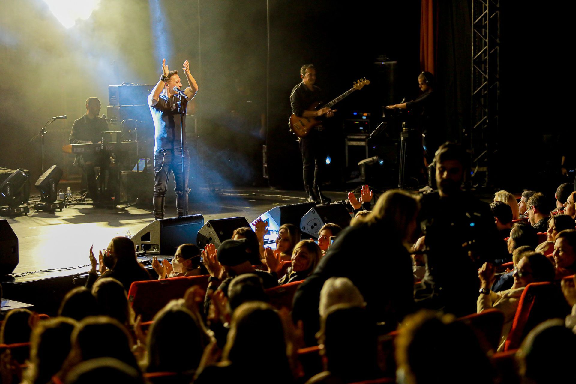 Um cantor se apresenta em um palco com pouca luz, acompanhada por uma banda, enquanto a plateia aplaude das cadeiras.