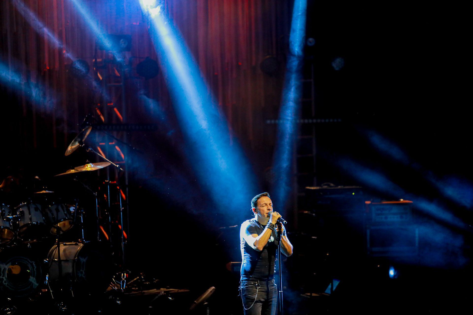 Homem cantando no palco sob holofotes azuis, com bateria ao fundo.