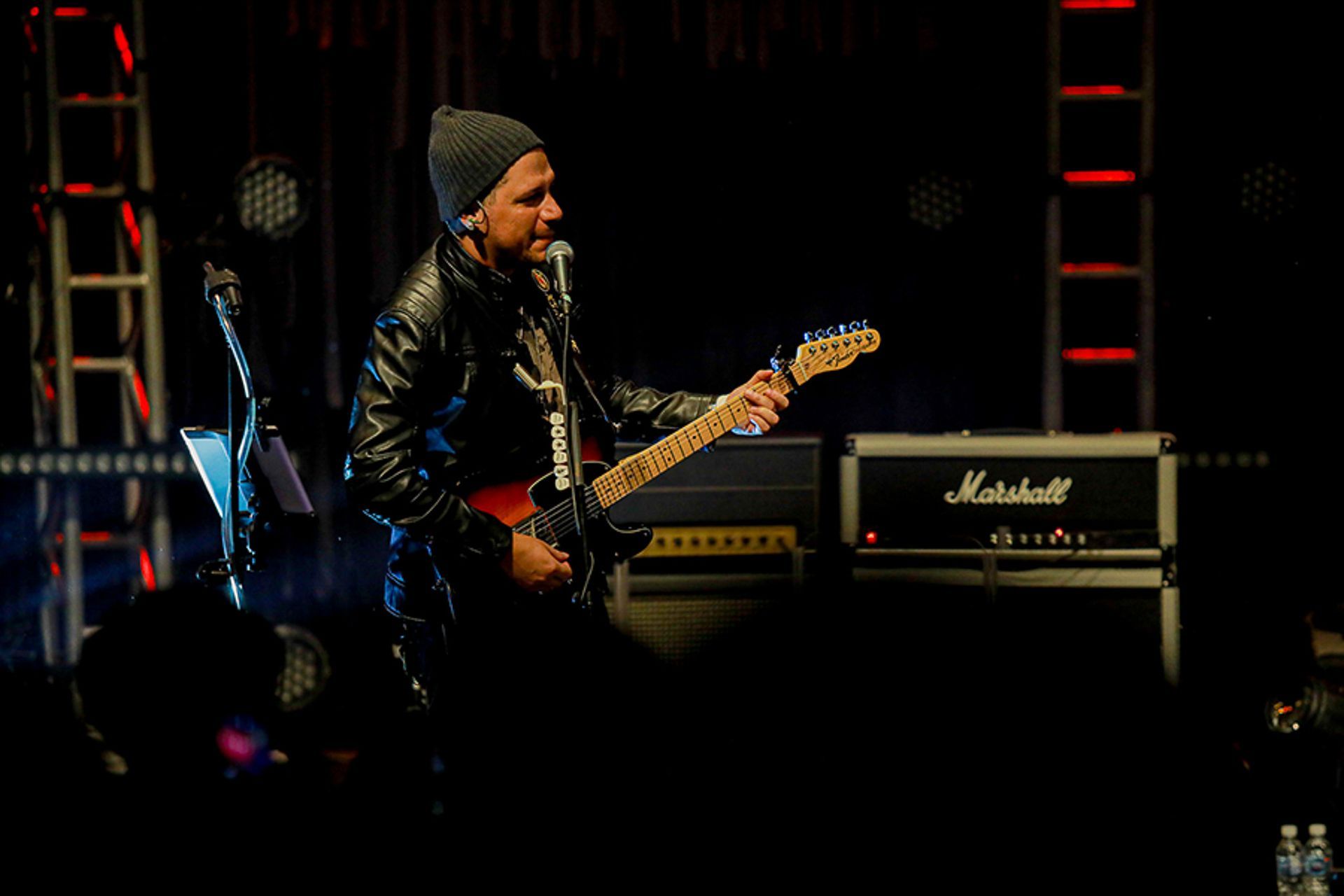 Uma pessoa usando gorro e jaqueta de couro toca guitarra no palco perto de um amplificador Marshall.