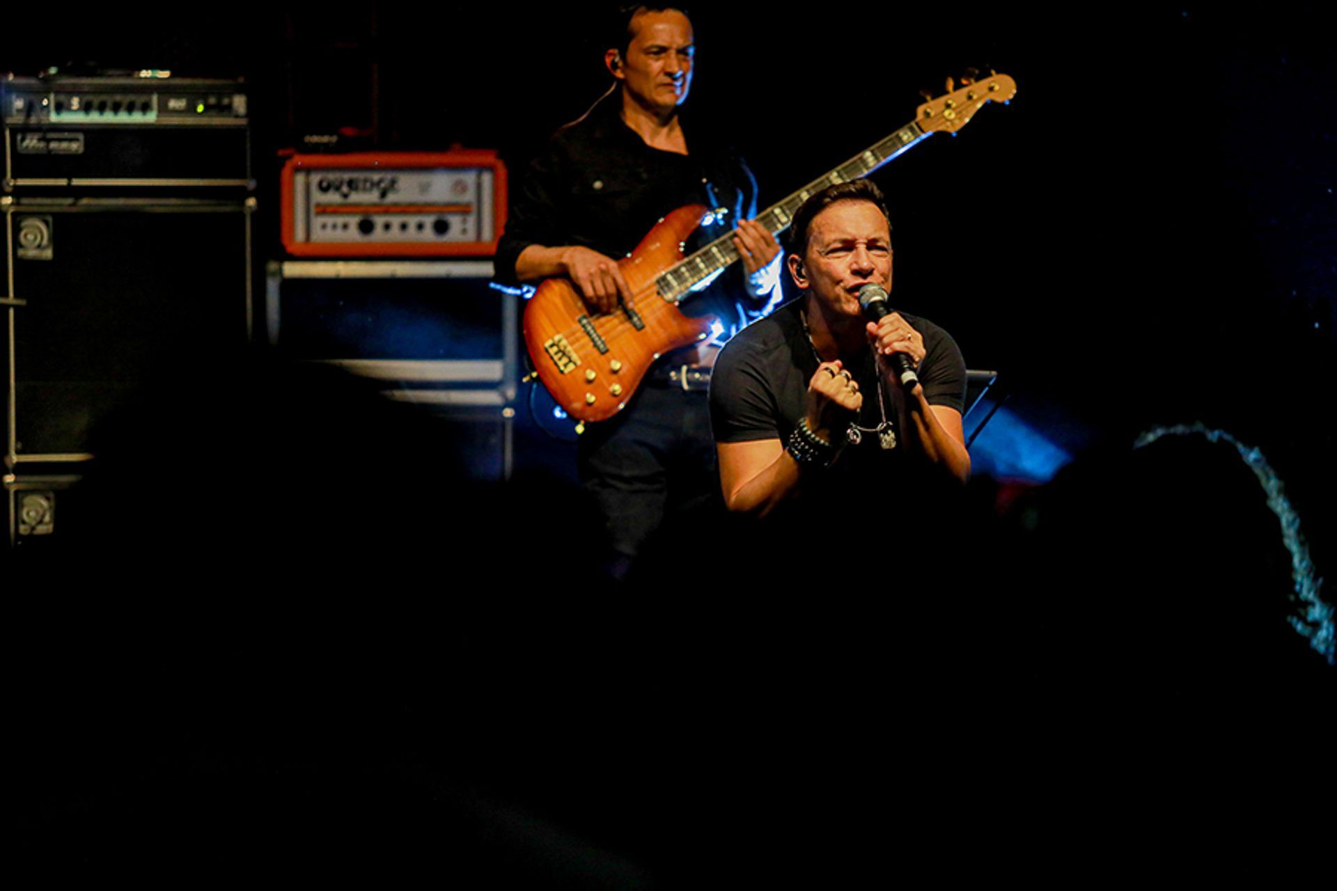 Um cantor se apresenta ao microfone enquanto um baixista toca um instrumento laranja no palco, em uma casa de shows escura.
