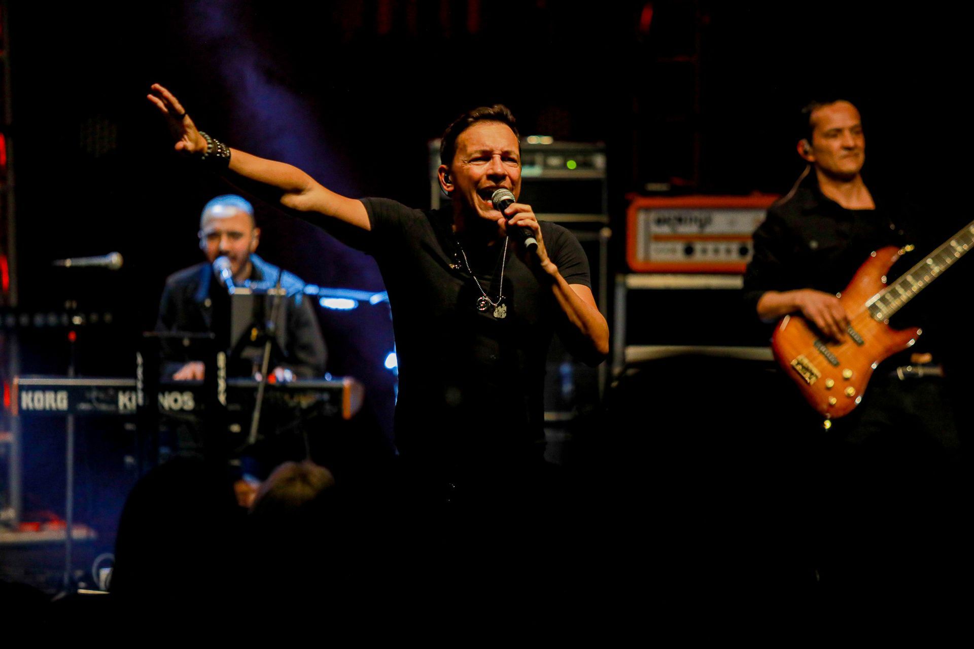 O vocalista principal se apresenta no palco com a mão estendida, acompanhado por um tecladista e um baixista.