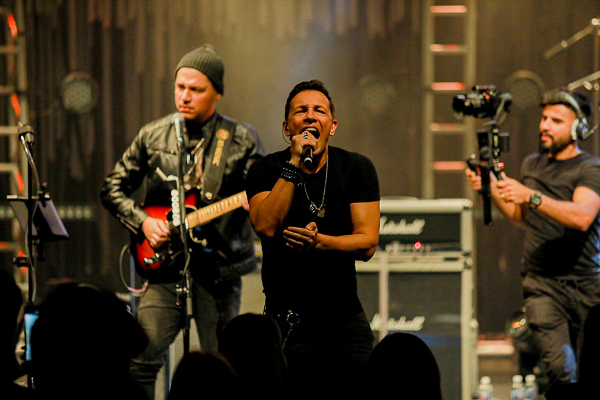 O vocalista principal se apresenta ao microfone no palco, acompanhado por um guitarrista e um operador de câmera.