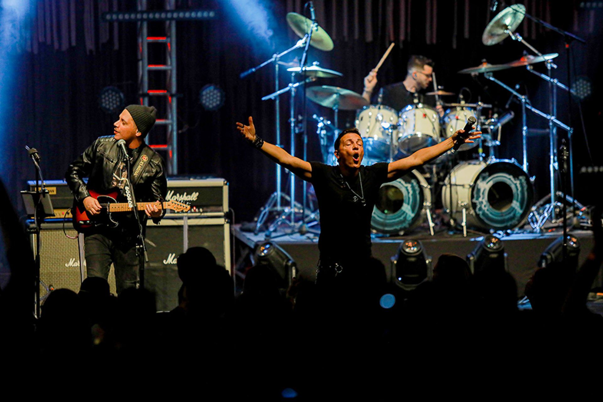 Banda se apresentando no palco com o guitarrista, o vocalista com os braços erguidos e o baterista.
