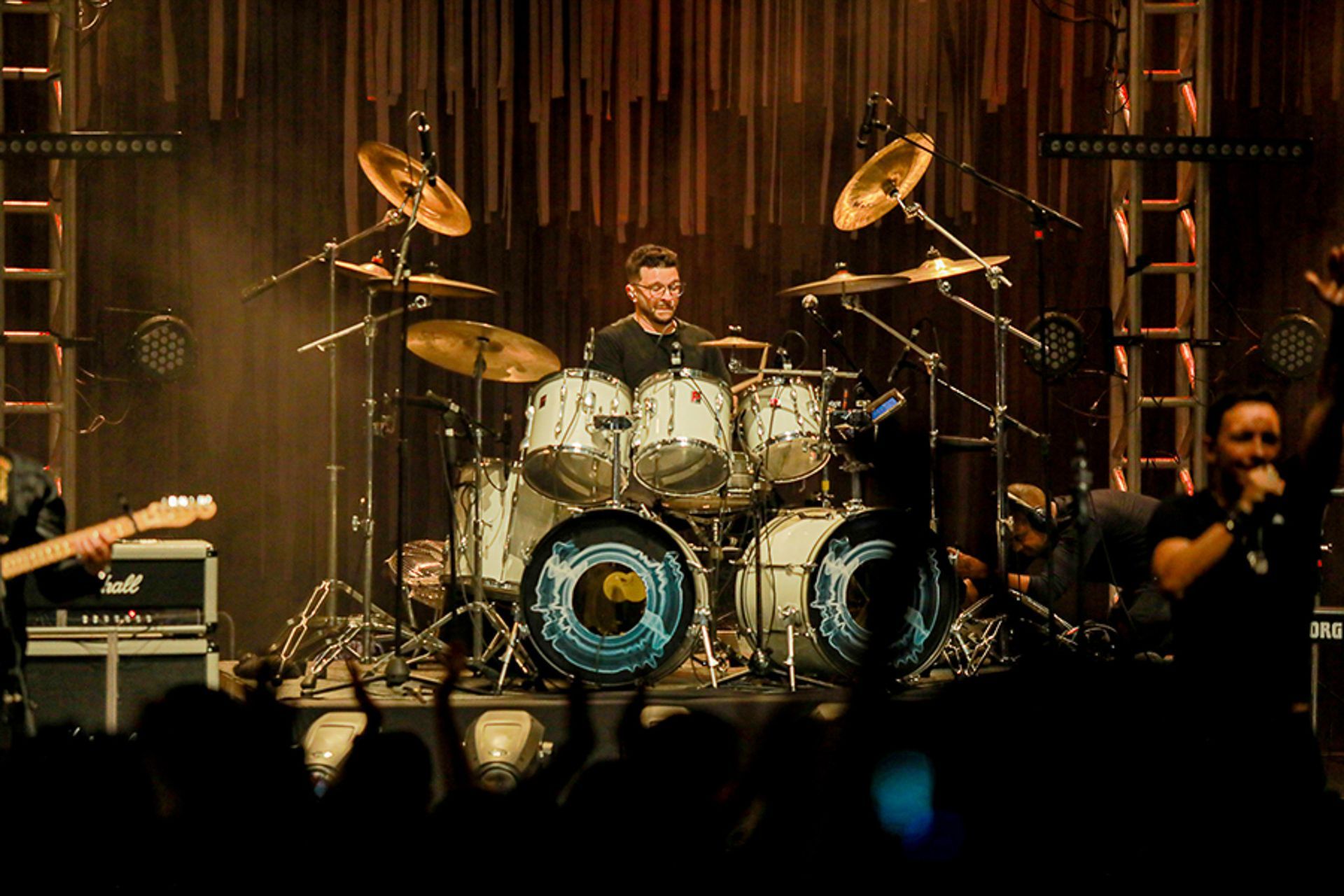 Um baterista toca no palco com uma banda, filmado da perspectiva da plateia em uma casa de shows escura.